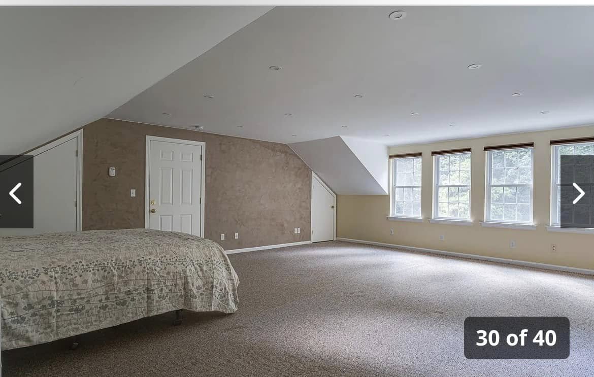 Spacious attic bedroom with sloped ceiling, beige walls, carpeted floor, four large windows, and a partially visible bed with a patterned bedspread.