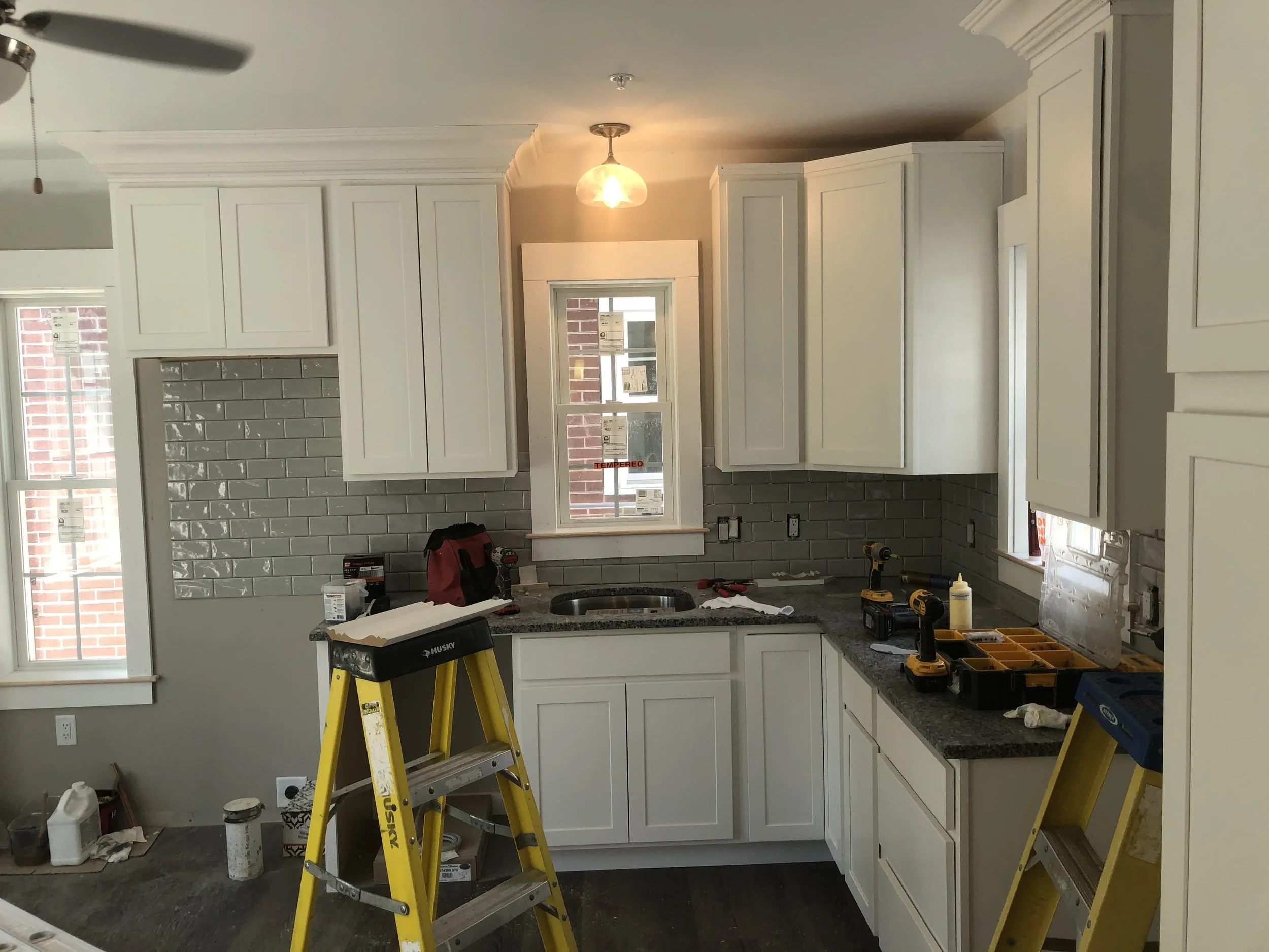 Kitchen under renovation with white cabinets, gray subway tile backsplash, and tools on the countertop, including drills and a toolbox, along with ladders.