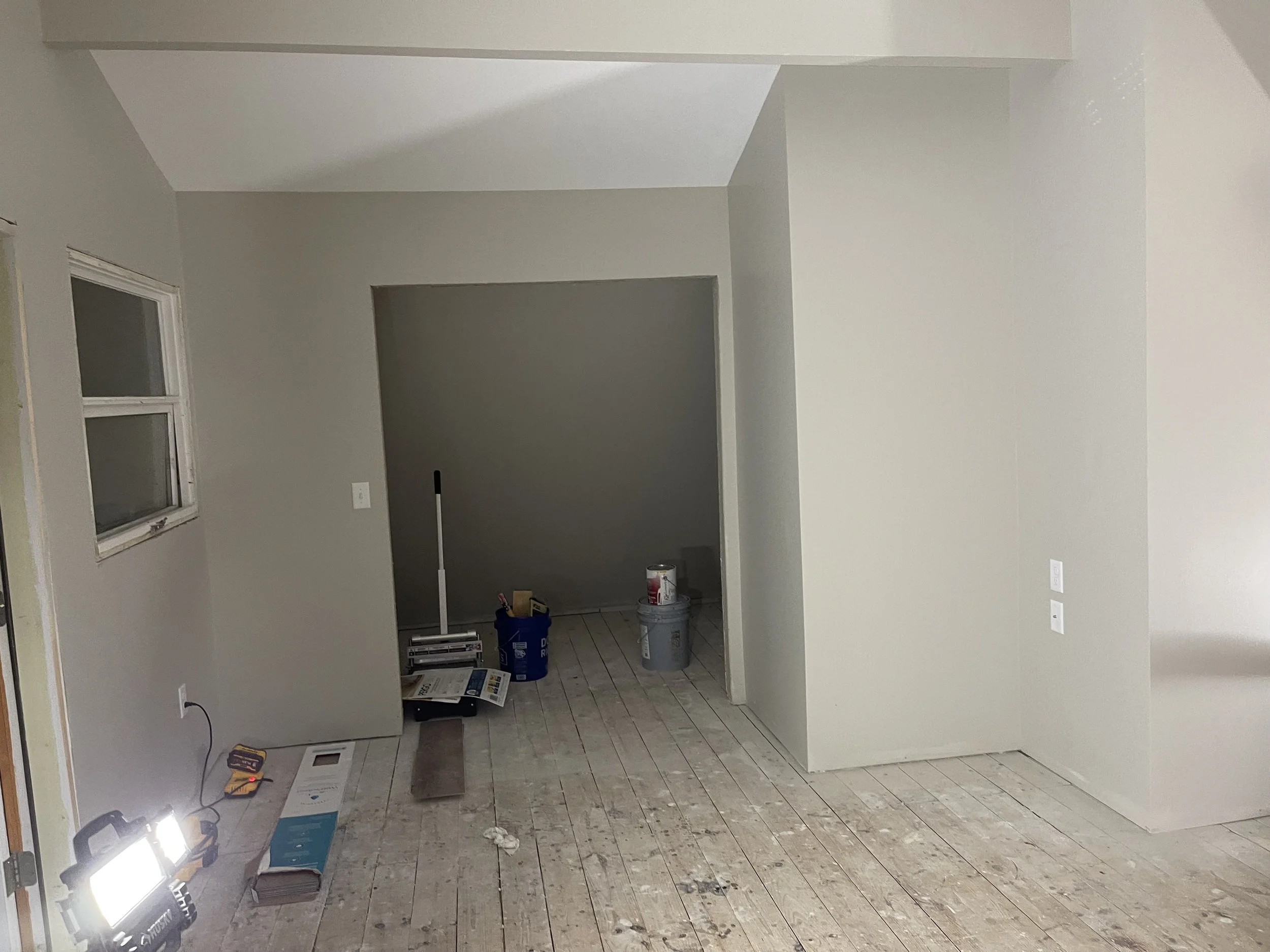 An interior room under construction with painted light-colored walls and exposed wooden floors. A small window is on the left, and the room opens into a smaller space or closet on the back wall. Construction tools and supplies, including a work light