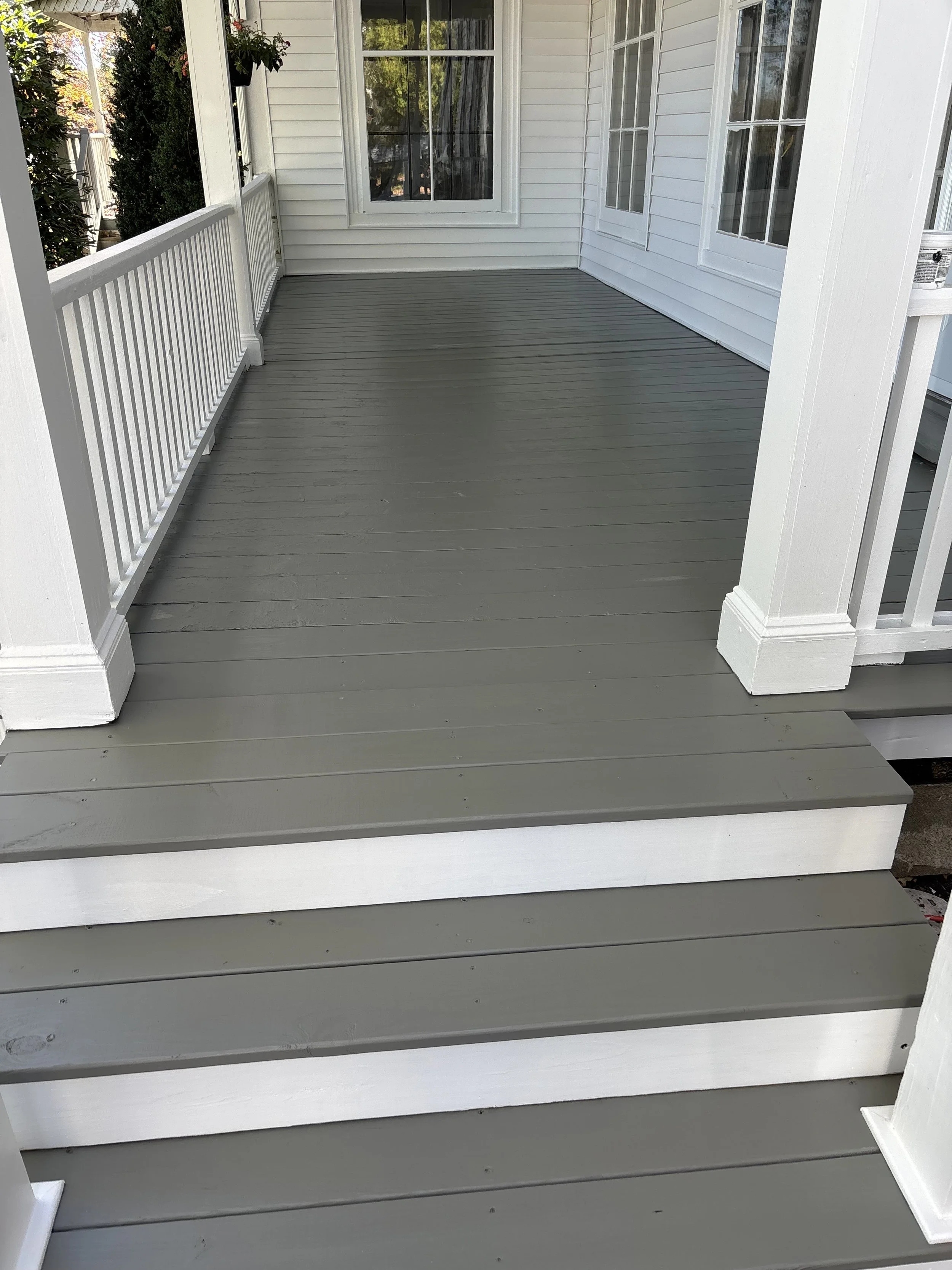 Front porch with gray wooden flooring, white railing, columns, and white house siding with large windows.