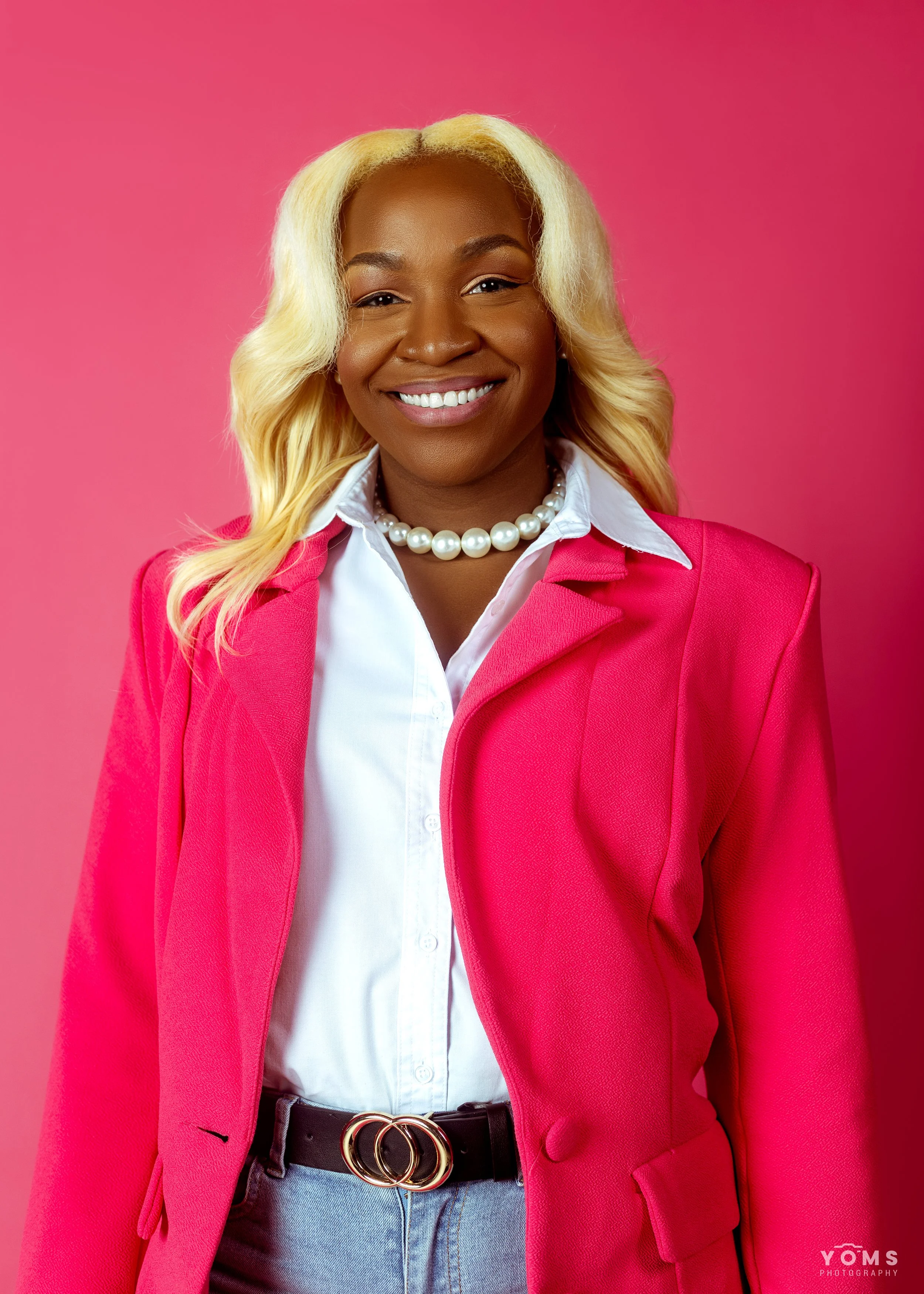 A woman smiling, wearing a white shirt, pink blazer, pearl necklace, black belt, and denim jeans standing against a pink background.