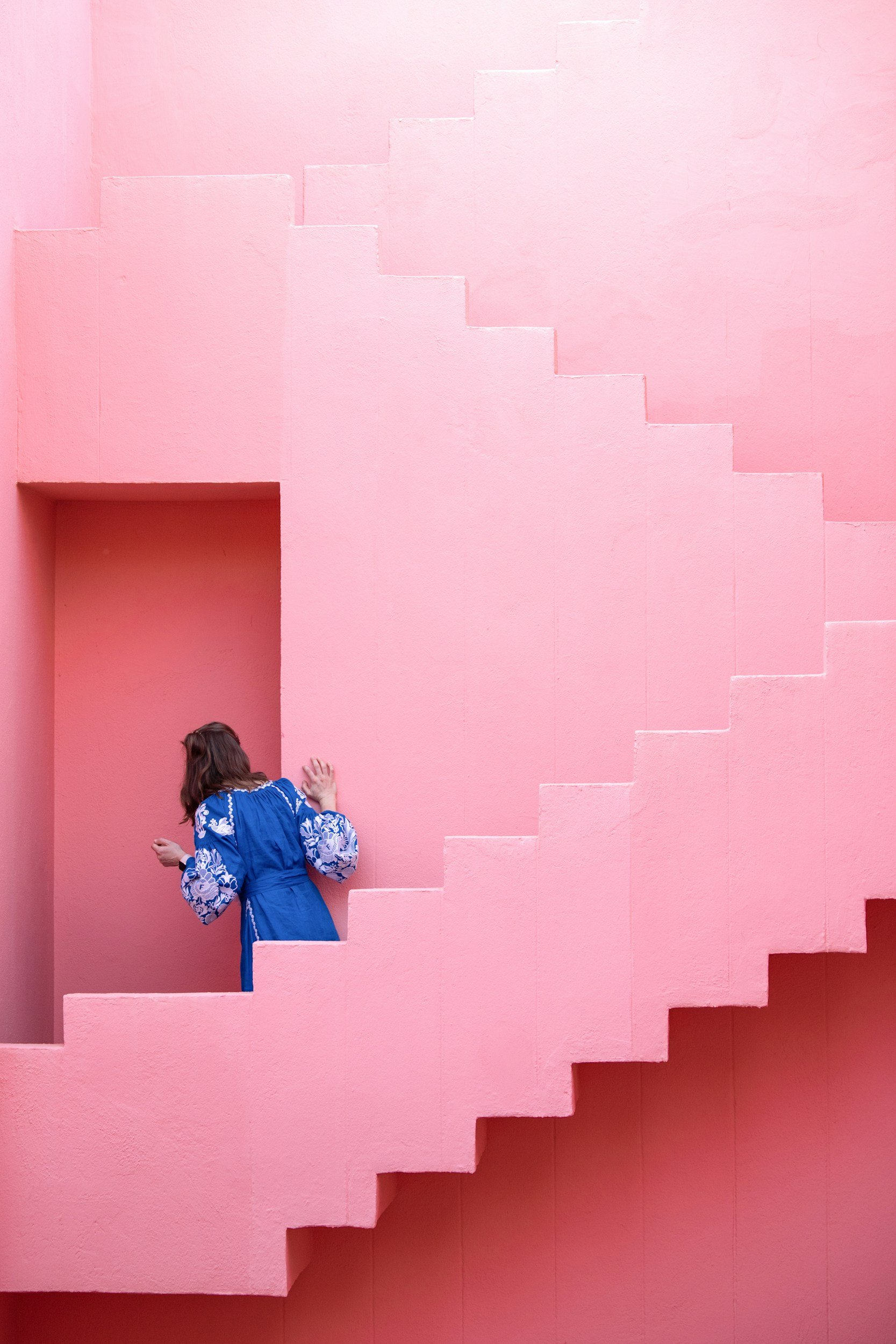 A woman in a blue dress with floral sleeves standing at the bottom of a pink staircase wall, her back facing the camera.