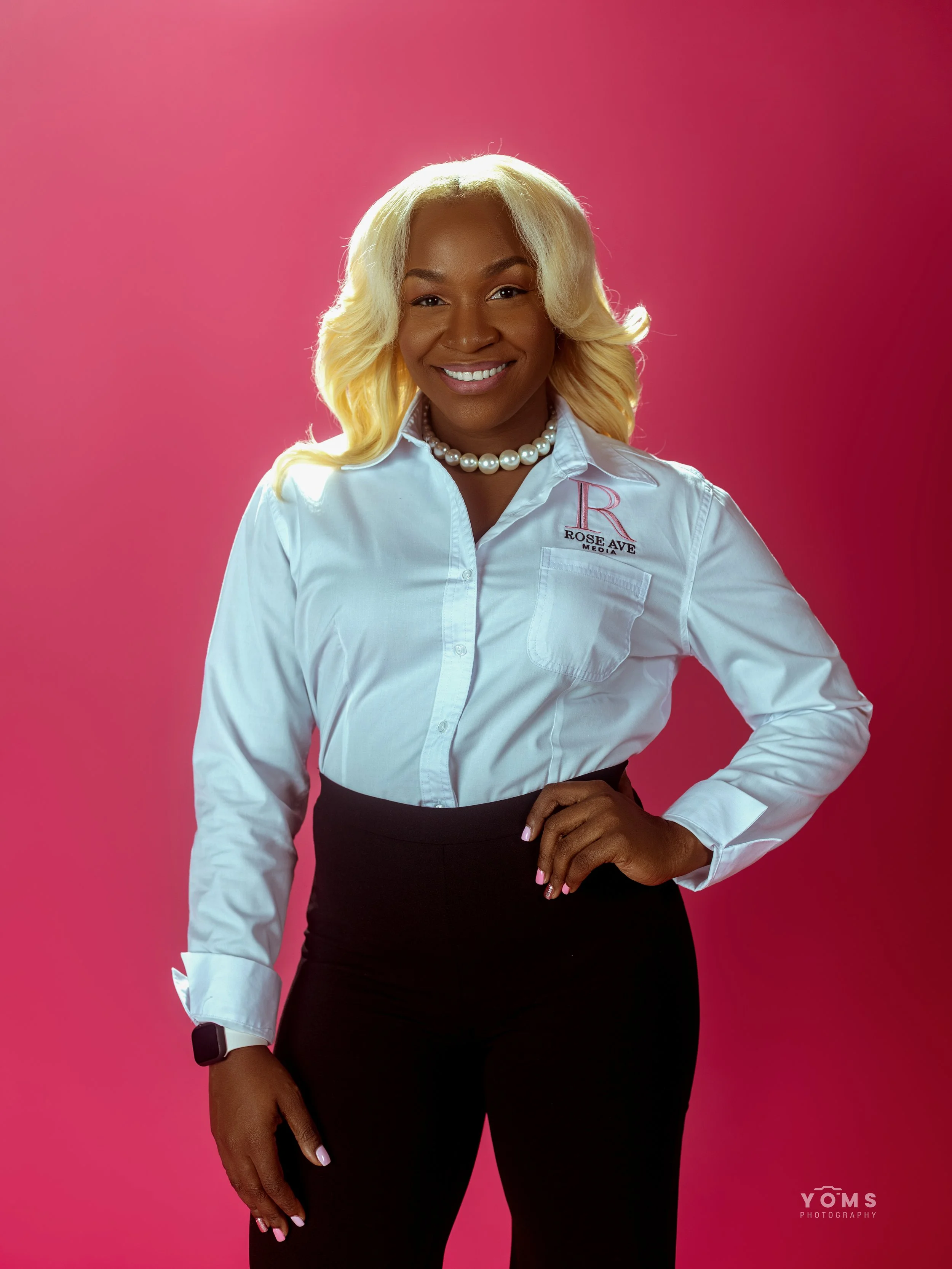 Professional woman with blonde curly hair, wearing a white shirt with the logo 'Rose Ave Media,' black pants, a pearl necklace, and a watch, posing with one hand on her hip against a pink background.
