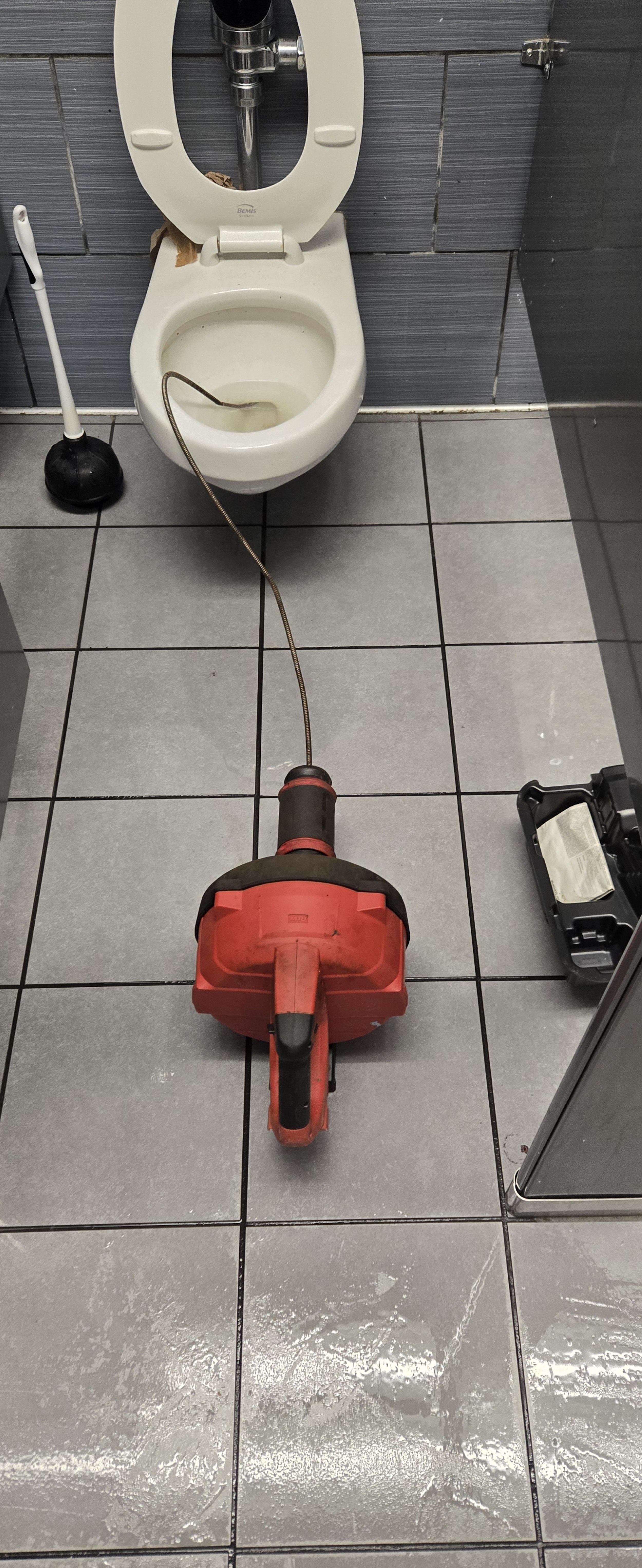 A toilet in a bathroom with a drain snake machine in front of it. The toilet is closed, and there is a plunger to the left. The tile floor is grey and shiny.