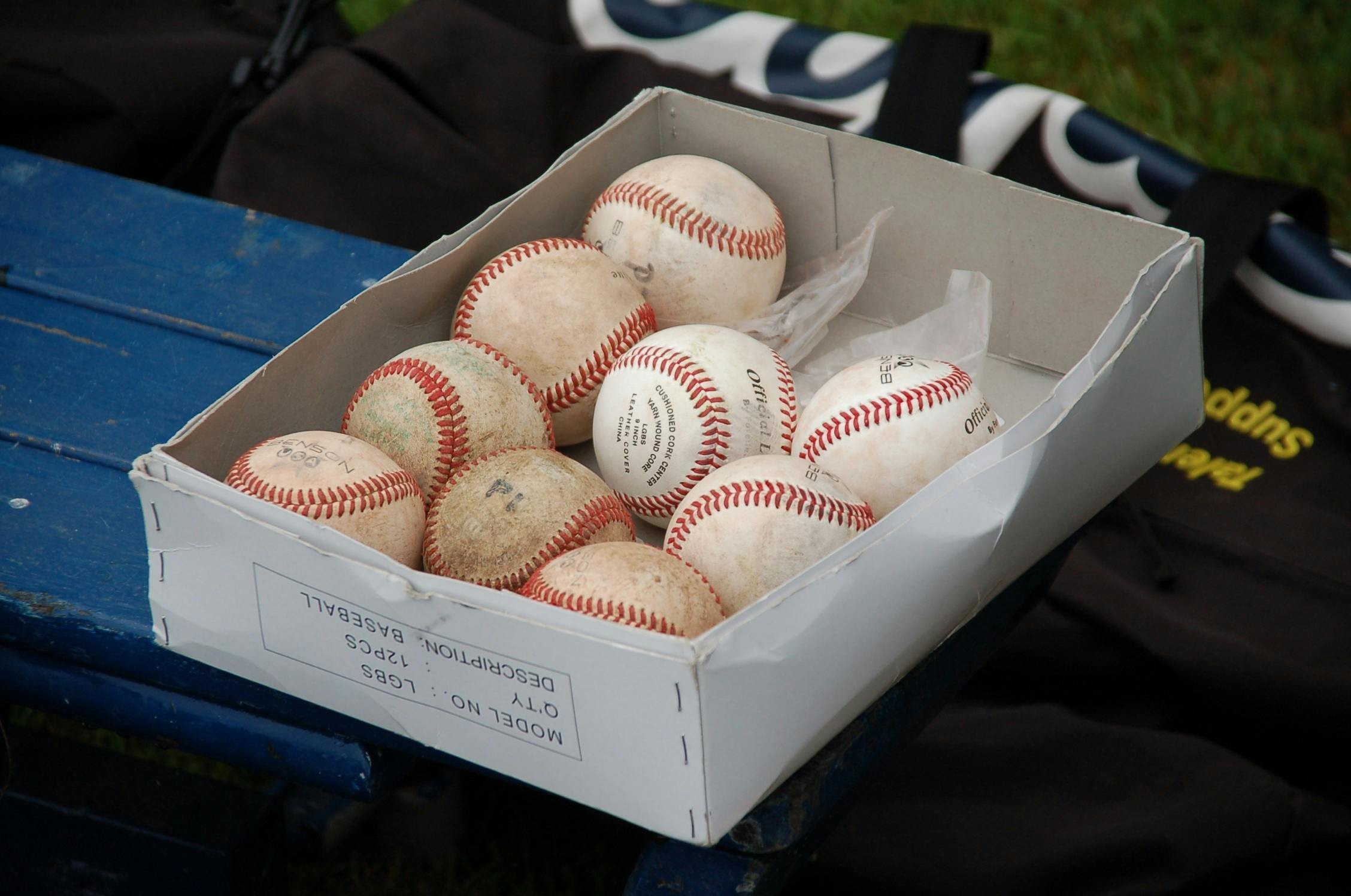 Old and dirty baseballs in a white cardboard box on a blue surface, with some grass and objects in the background.