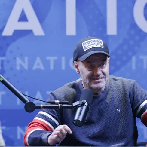 A man in a navy blue sweatshirt, wearing a baseball cap, speaking into a microphone during a podcast or radio show with a blue background.