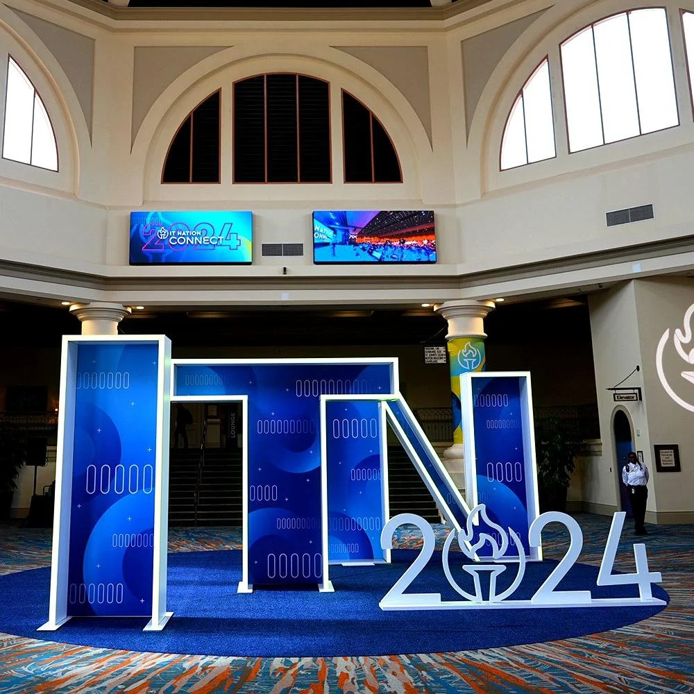 Indoor conference center with large blue and white 2024 sign featuring a torch logo, with four blue panels, two digital screens above showing conference information, and a patterned carpet.