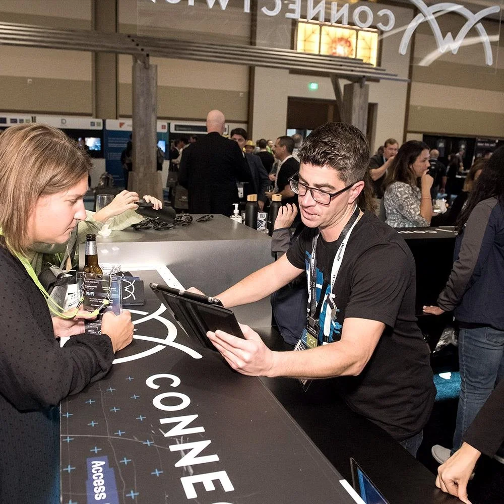 A man and a woman at a conference registration desk, with the man showing something on a tablet and the woman holding a badge, surrounded by other attendees in a busy event setting.