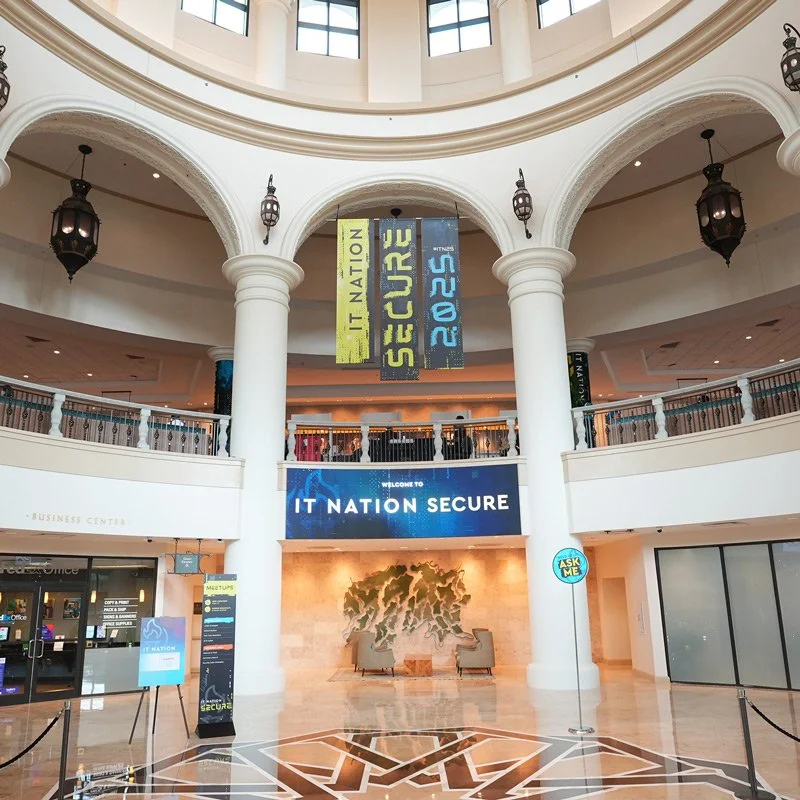 Interior of a large shopping mall or conference center with a high, domed ceiling, white classical columns, and hanging lantern-style lights. There are banners hanging that read 'IT NATION SECURE 2023'. There is a reception area with a sign that says 'WELCOME TO IT NATION SECURE' and two chairs facing each other. There is an 'ASK ME' sign on a stand near the entrance to a store or office.