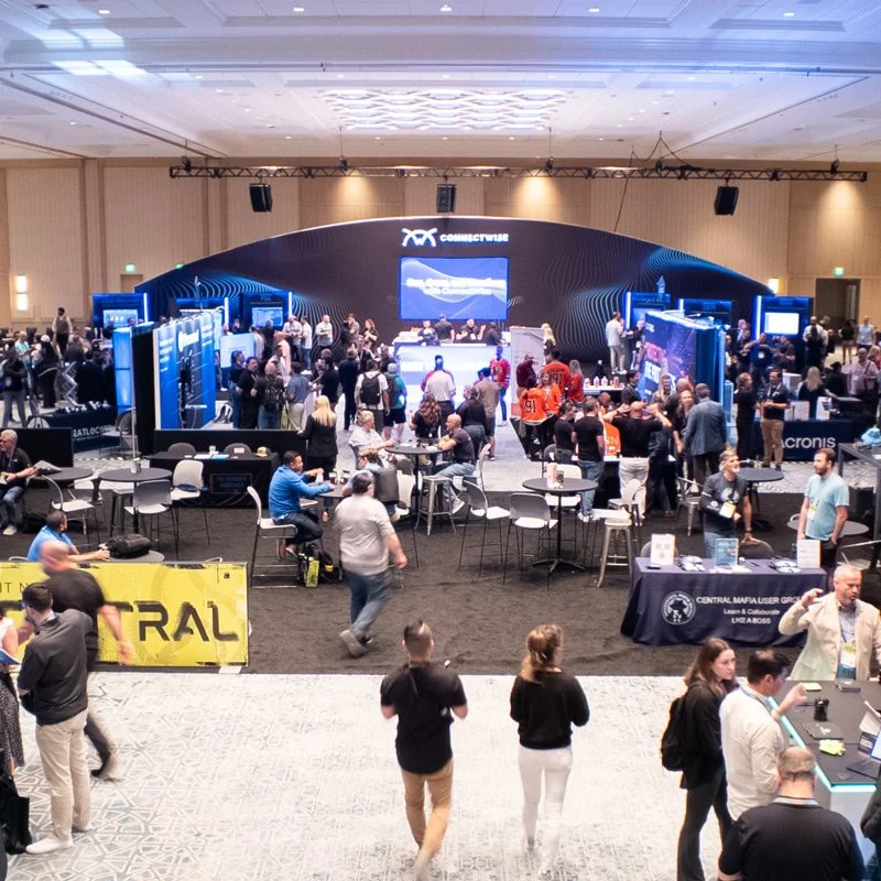 Large tech conference with numerous attendees walking and sitting around exhibition booths and a stage with a large screen in a ballroom setting.
