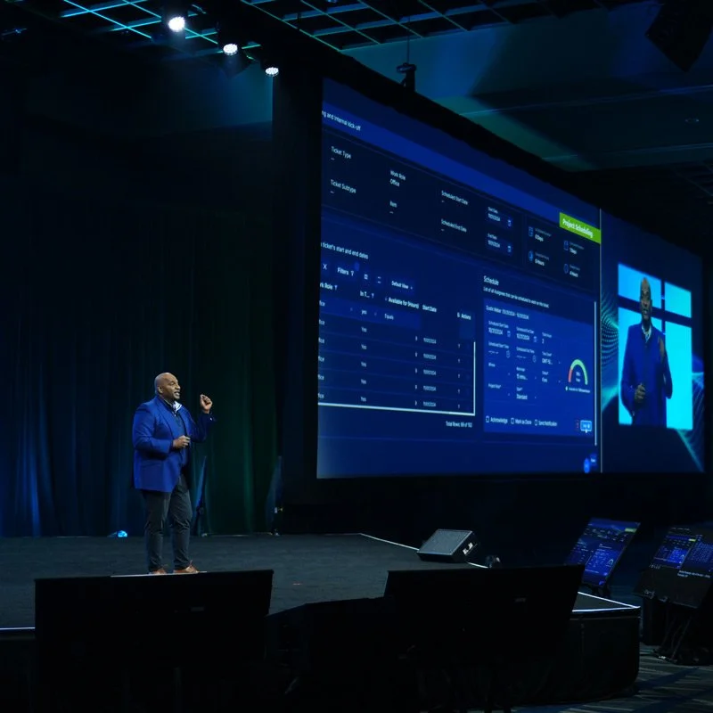 A man in a blue blazer presenting on stage with a large screen behind him displaying a software interface, during a conference or event.