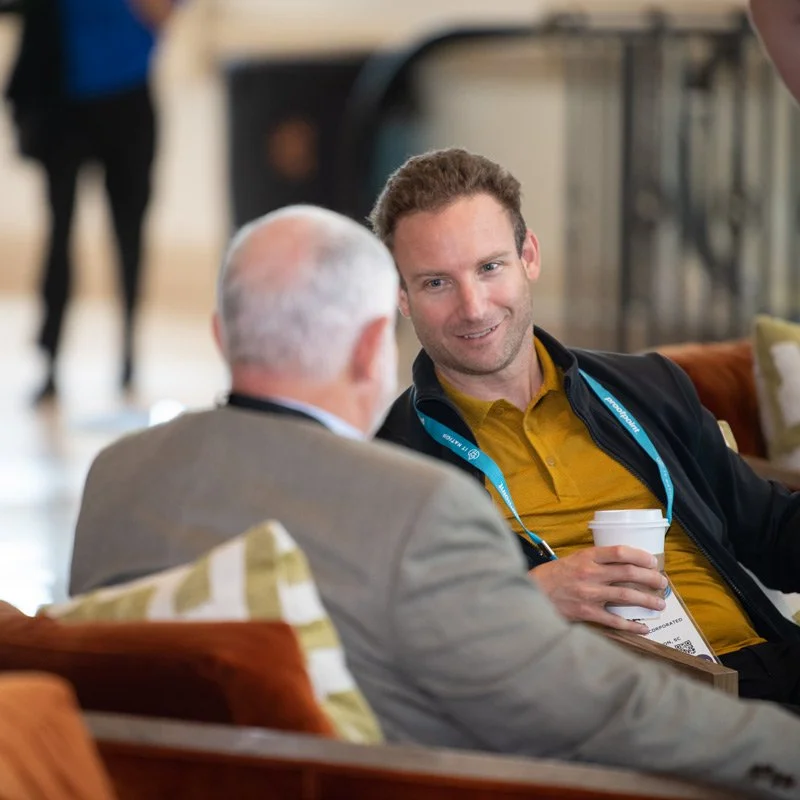 Two men seated and engaging in conversation in a hotel lobby or conference area. One is an older man with white hair, seen from behind, wearing a gray blazer. The other is a younger man with short curly hair, holding a white coffee cup, wearing a yellow shirt and black jacket with a conference badge around his neck.