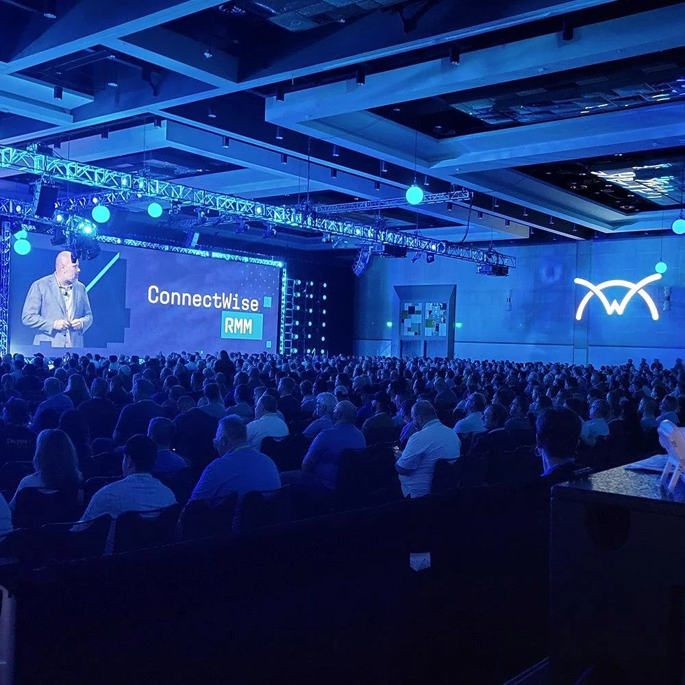 Large conference room filled with attendees watching a presenter on stage with large screens displaying 'ConnectWise RMM' and a logo on the wall.