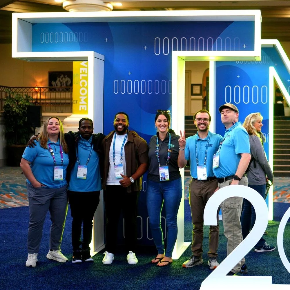 Group of six people standing together in front of a large blue and white abstract background at a conference, smiling and posing for the photo, some making peace signs.