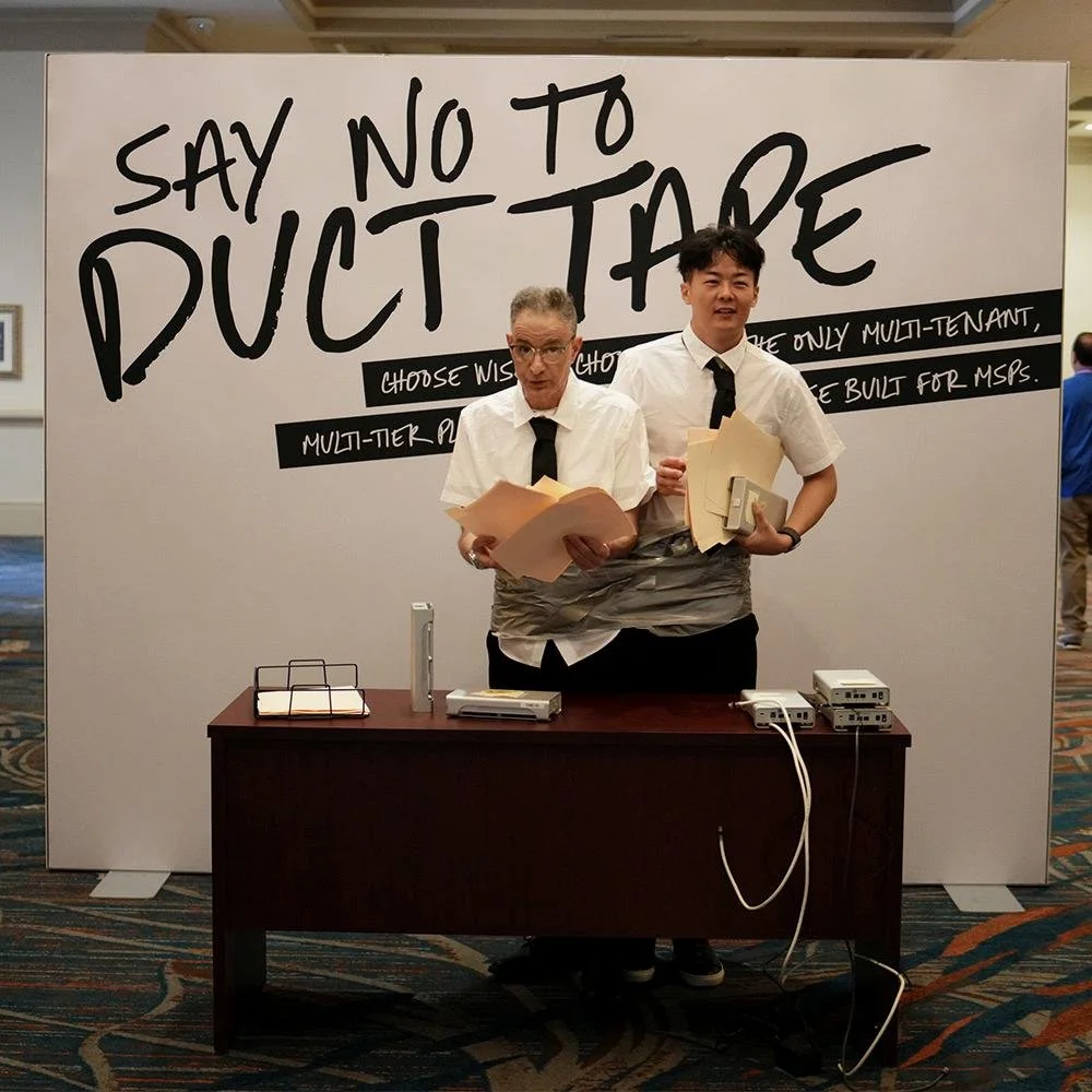Two men dressed as office workers with white shirts and black ties standing behind a desk, holding papers, in front of a large sign that says 'Say No to Duct Tape'.