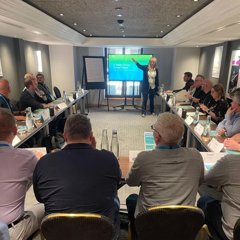 Business meeting or conference in a modern conference room with a female presenter at the front pointing to a digital screen, surrounded by attendees seated in a U-shape arrangement.