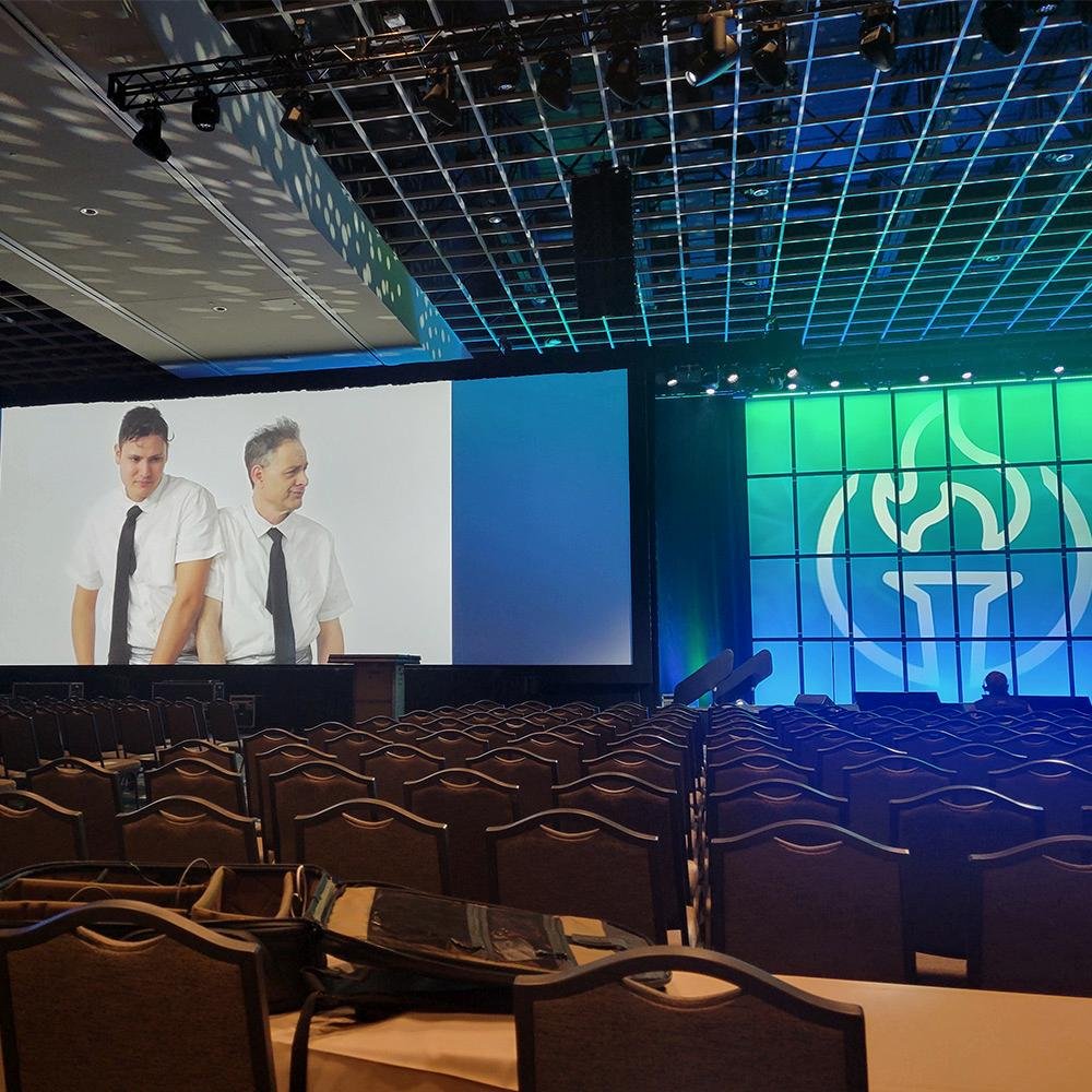 An empty large conference or event hall with rows of chairs, a big screen showing two men in white shirts and black ties, and a colorful backdrop with a flame logo.