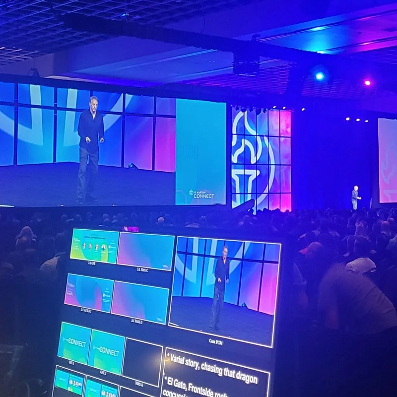 A large conference hall with a speaker on stage, presenting to an audience. Multiple screens display various slides and visual content related to the presentation.