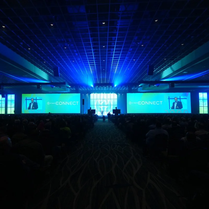 Large conference room filled with attendees, stage with multiple large screens displaying a speaker at the 2018 NATI CONNECT conference, blue lighting.