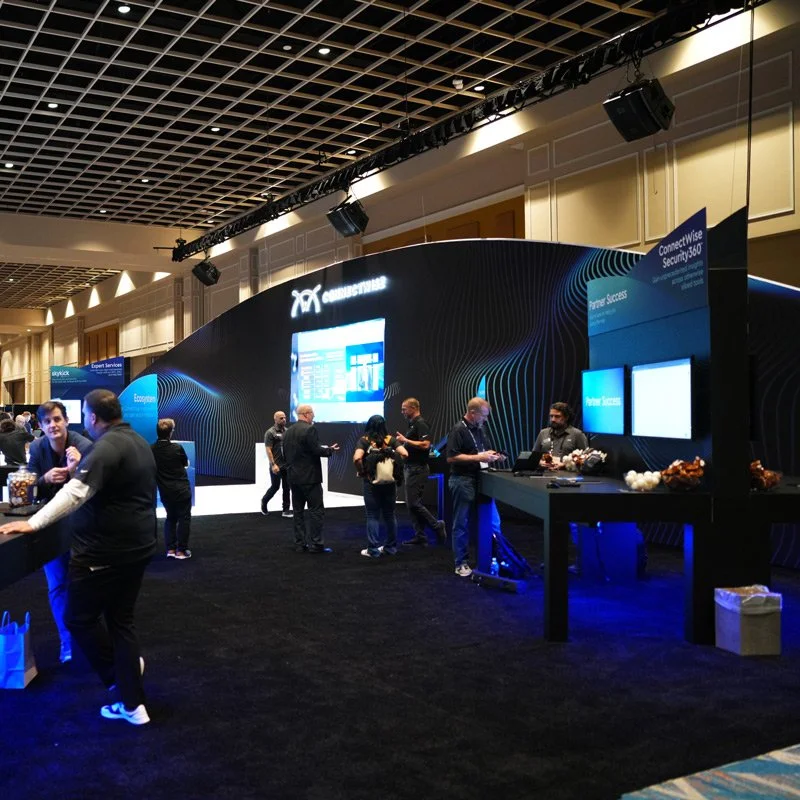 People visiting a technology exhibition booth with digital displays and promotional material, some engaging with staff, under a high black grid ceiling with lights.