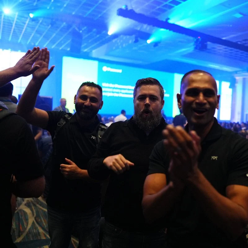 Three men at a conference or event, smiling and cheering, with a large blue screen and technology setup in the background.