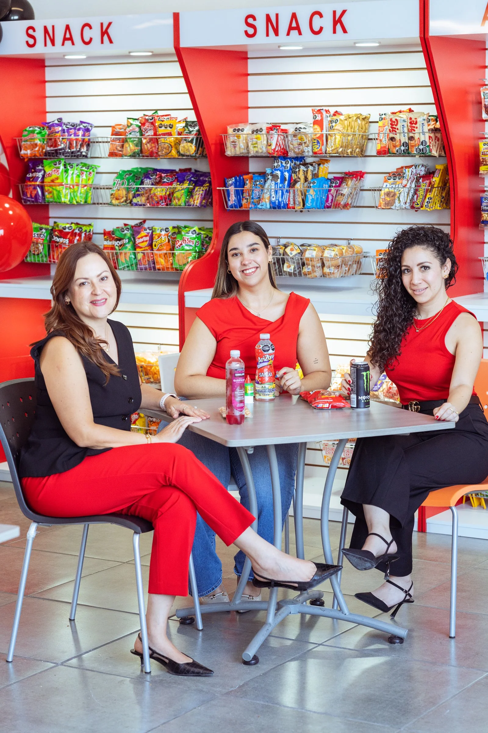 Tres mujeres sentadas en una mesa en una tienda de snacks, con estantes llenos de diferentes tipos de snacks en el fondo