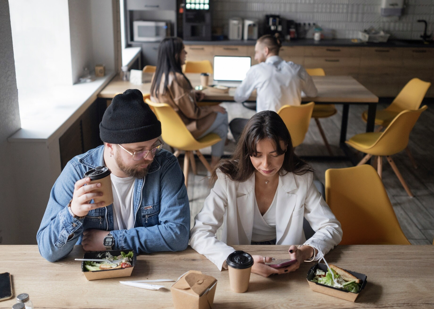 Dos personas sentadas en una mesa en un café, uno con una taza y la otra con un teléfono móvil, con comida para llevar y bebidas, y otras personas trabajando en el fondo.