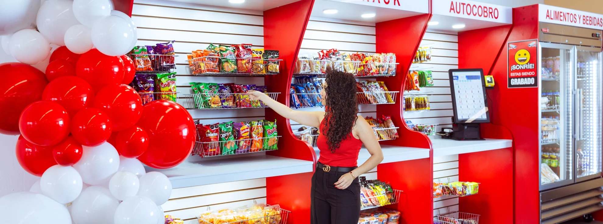Mujer comprando snacks en una tienda o supermercado, con decoración de globos rojos y blancos.