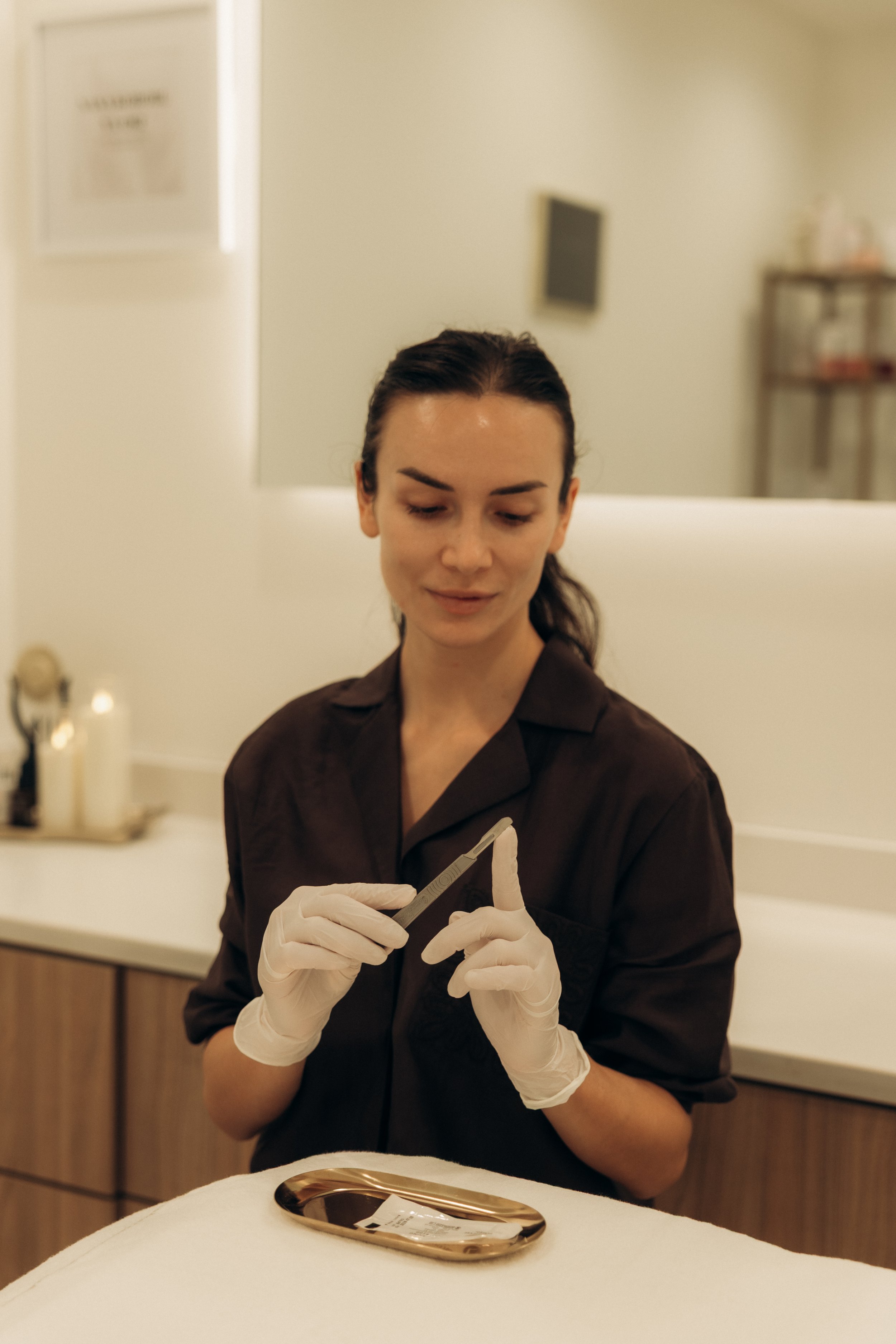 A woman wearing white gloves is holding a pair of tweezers, likely during a cosmetic or dermatological procedure indoors.