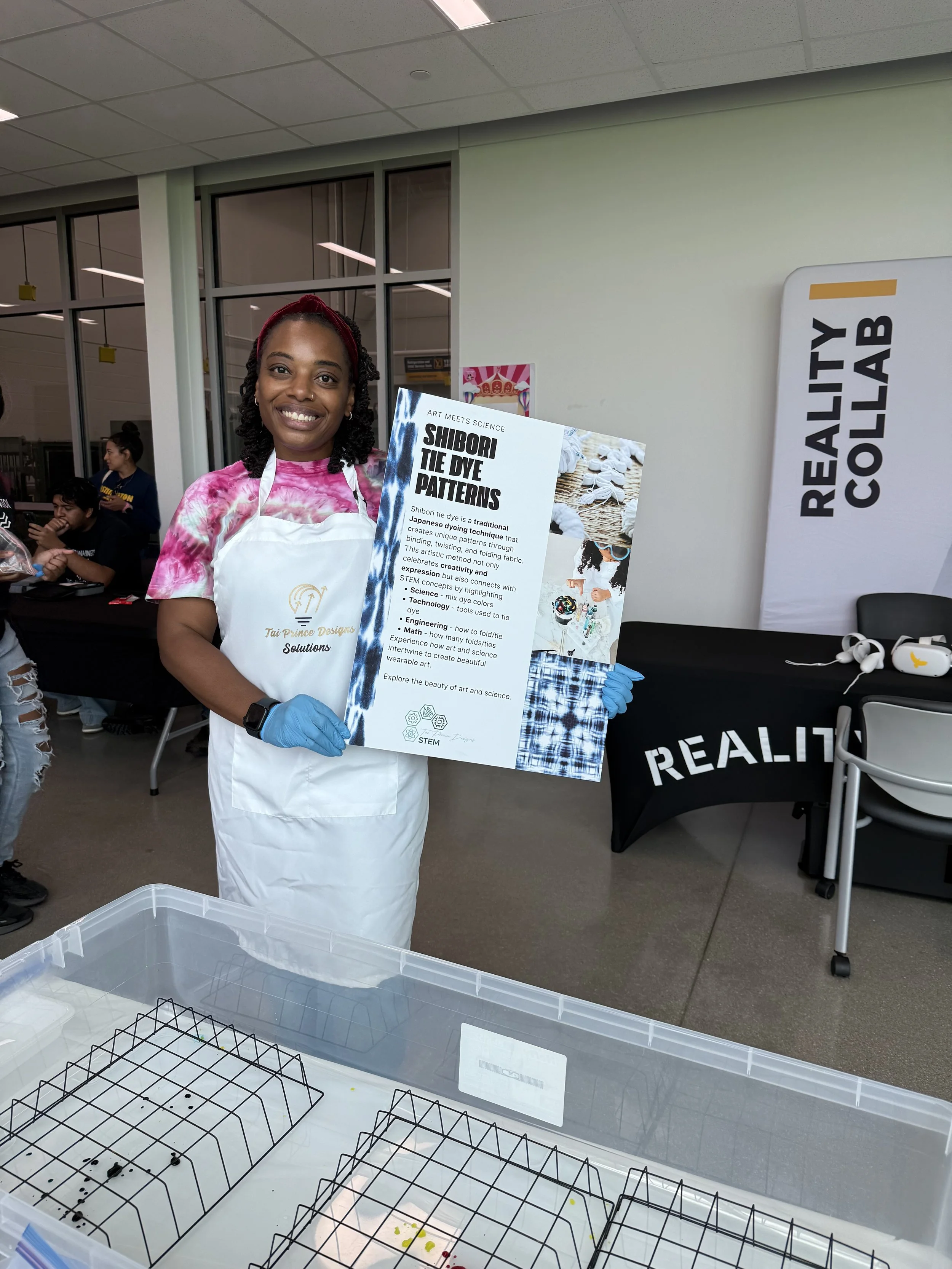 A woman smiling and holding a sign about Shibori tie dye patterns at a science art event, wearing a pink tie dye shirt, white apron, and blue gloves, in a room with other people and a table with scientific displays.