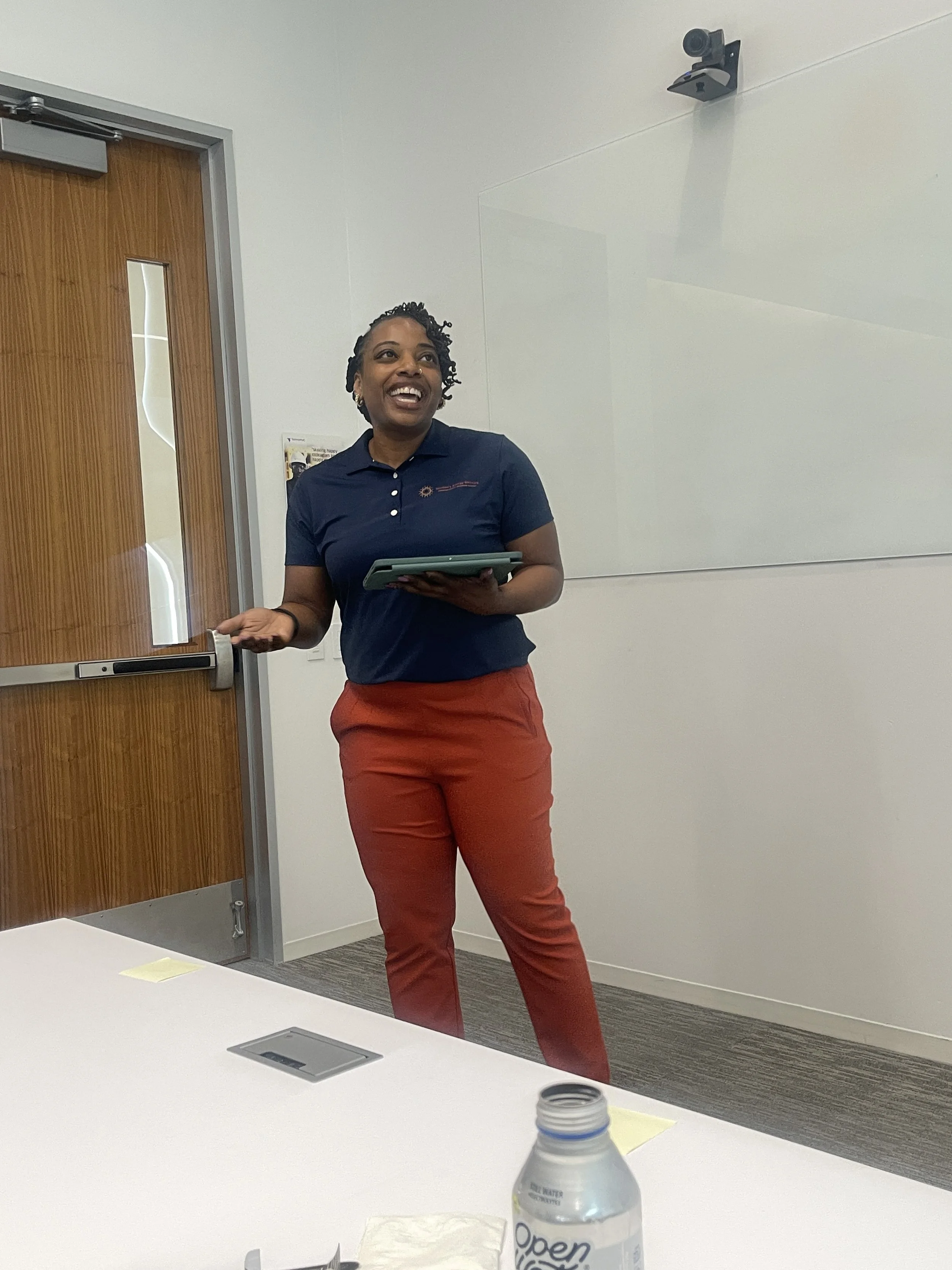 A woman standing in a conference room giving a presentation with a smile, holding a tablet in her hand, near a whiteboard and a door.