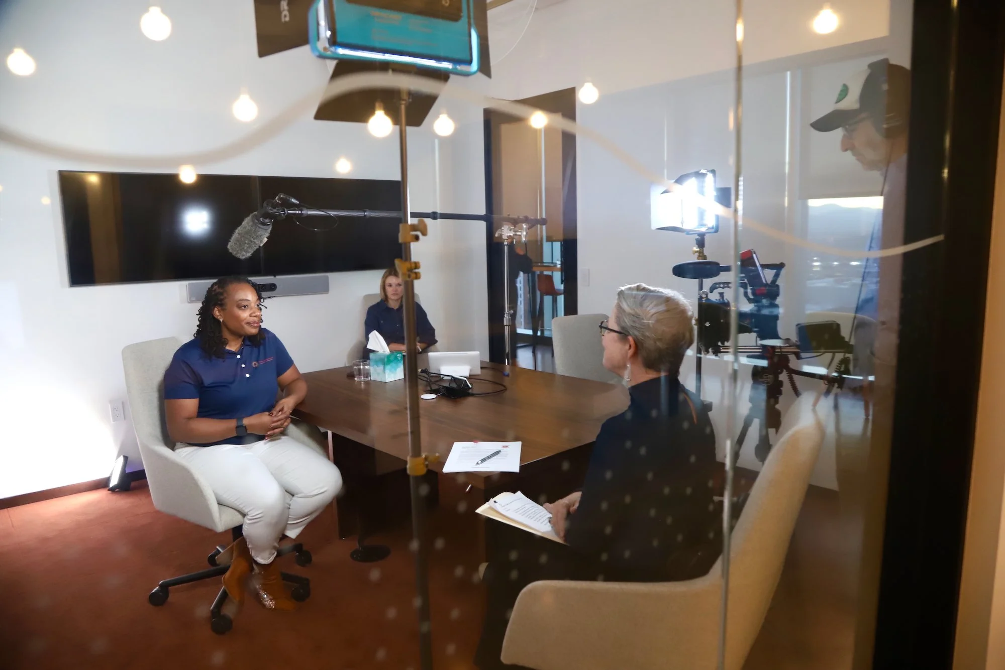 A woman in a blue shirt being interviewed in a conference room with camera and lighting equipment set up.