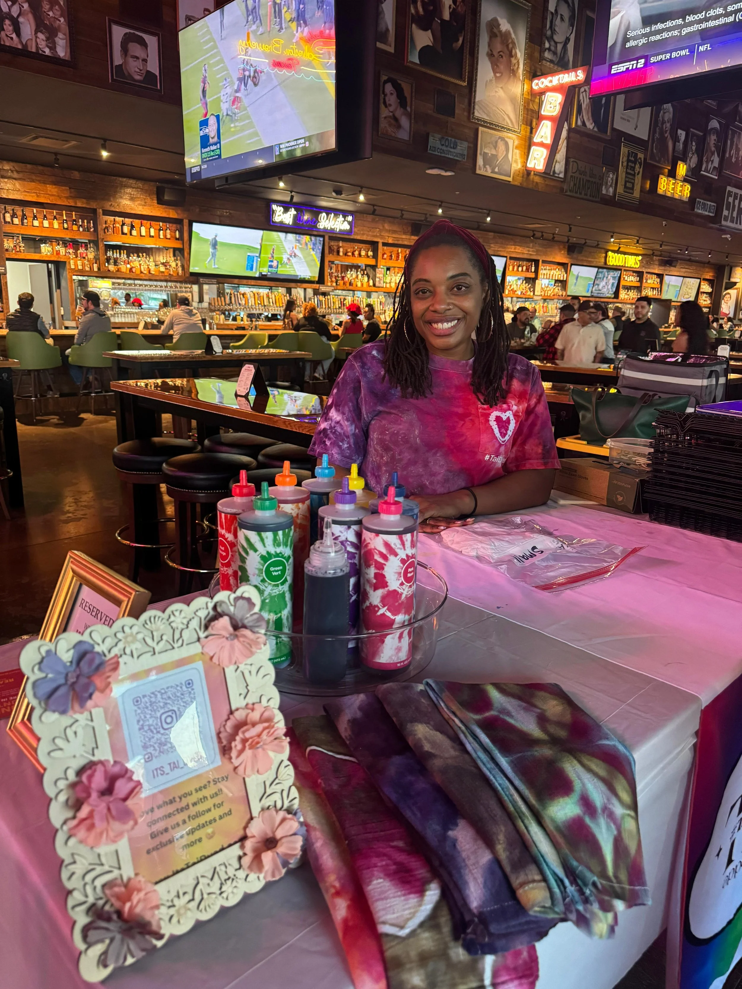 A woman with dreadlocks smiling at a table in a busy bar or restaurant decorated with colorful graphics and neon signs. The table has tie-dye bottles, folded shirts, and a sign with a QR code. People are seated in the background watching TV screens.