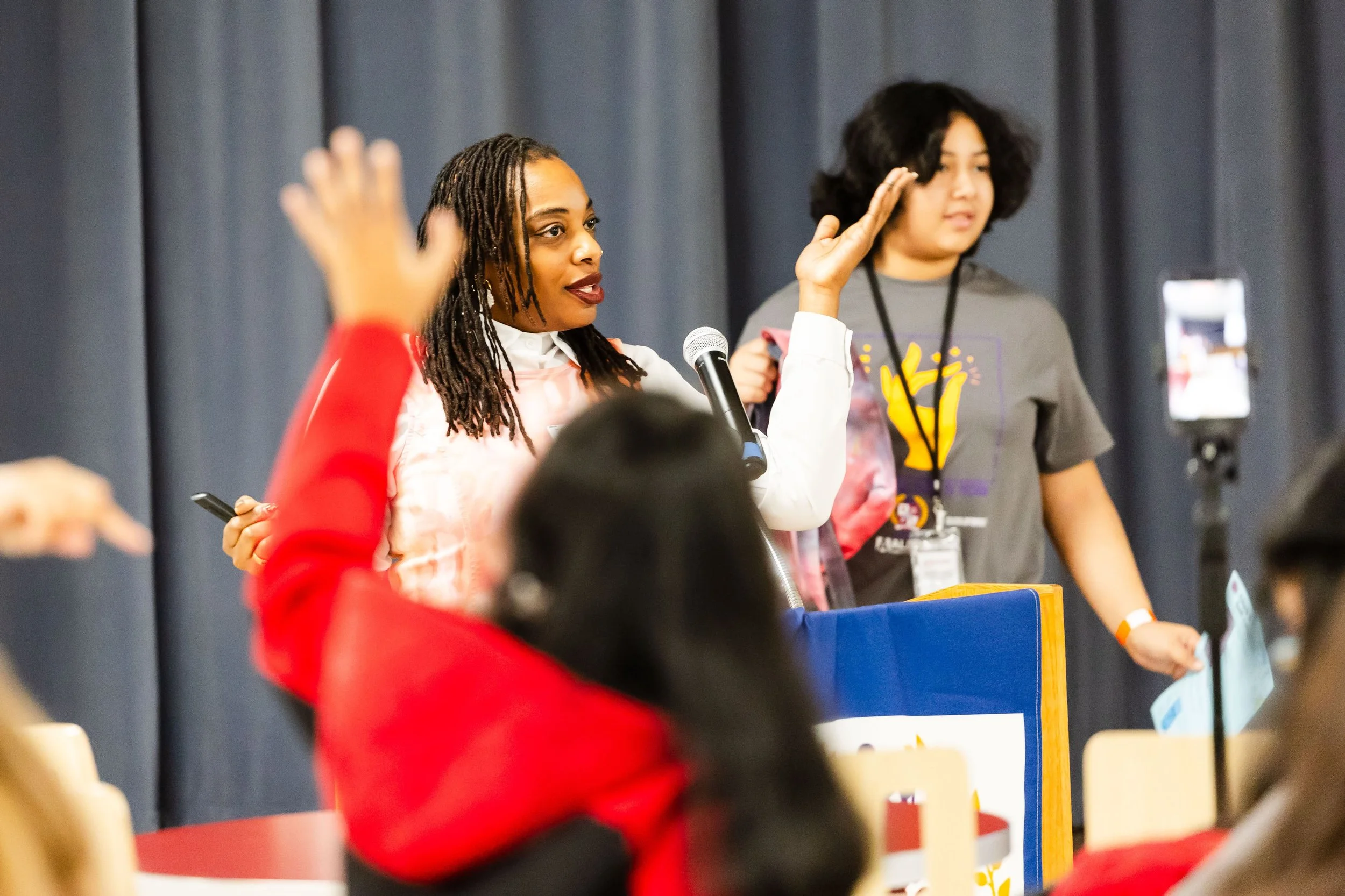 Two women speaking at a panel or conference, with one woman holding a microphone and gesturing with her hand, while the other woman stands next to her. Several people are visible sitting and recording or taking notes.