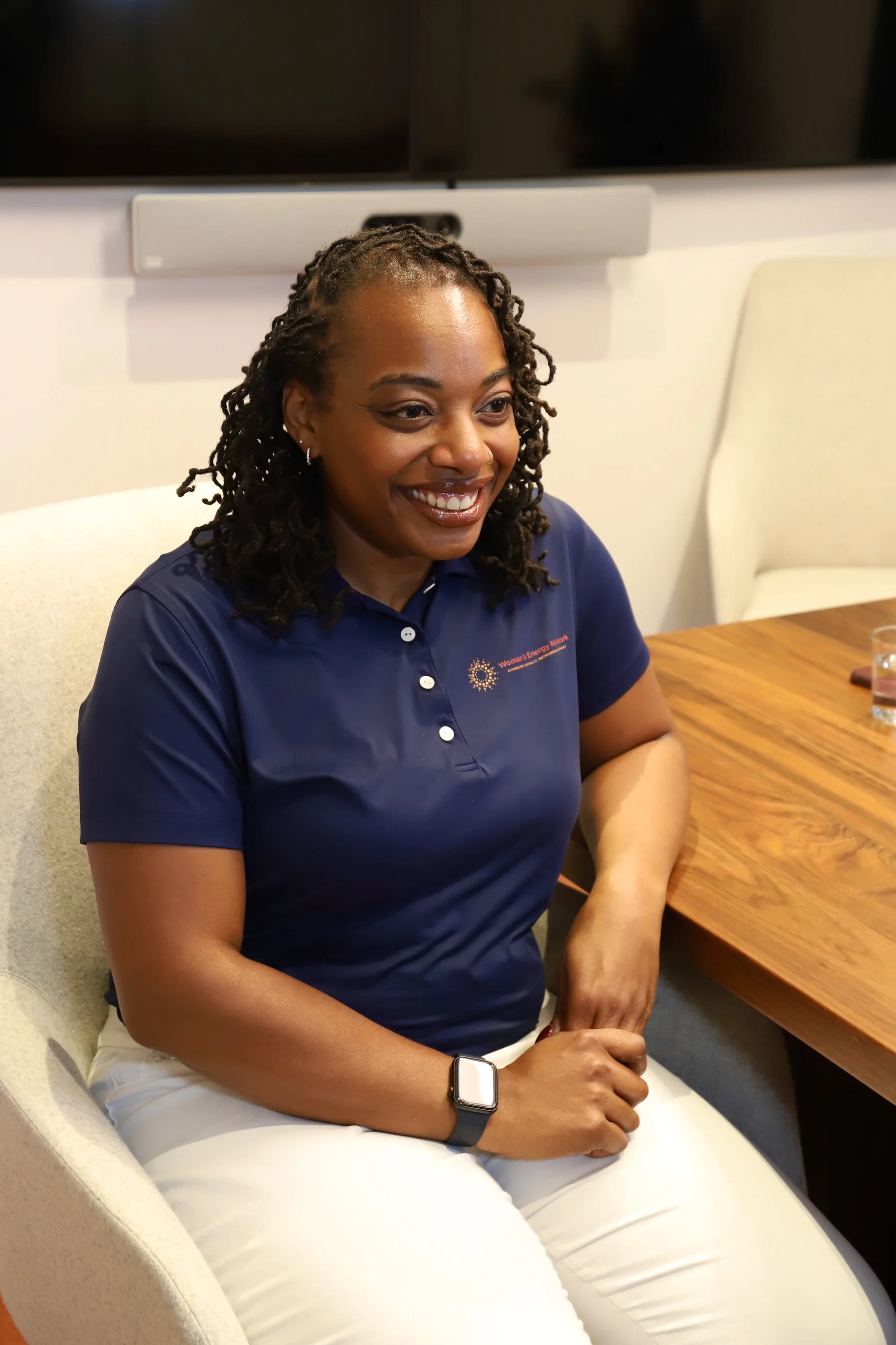 A smiling woman with dark curly hair, wearing a blue polo shirt, sitting at a wooden table with a glass of water, in a modern conference room.
