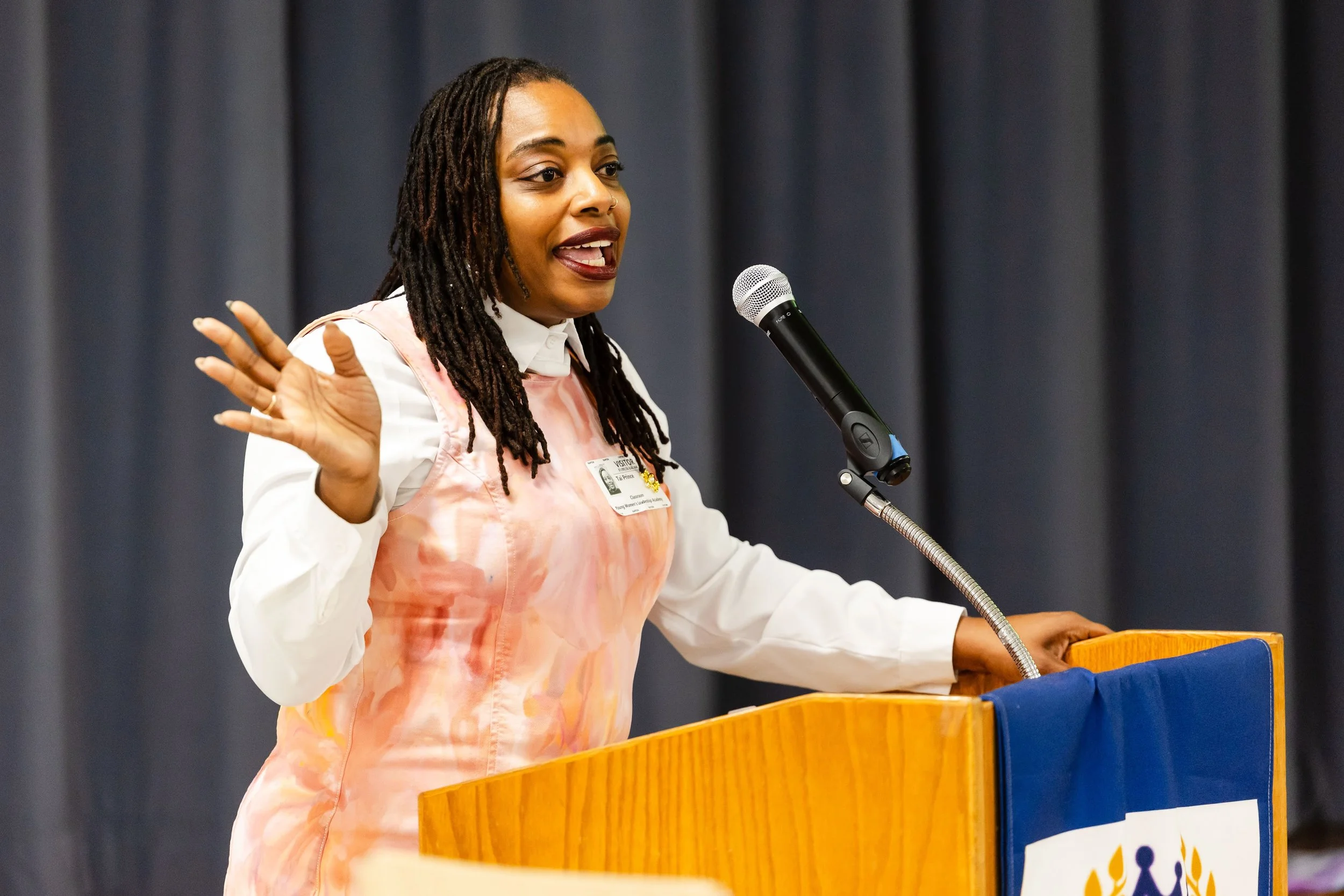 A woman with long dreadlocks speaking at a podium with a microphone in front of a dark curtain background. She is wearing a white shirt and a pastel pink apron, gesturing with her left hand.