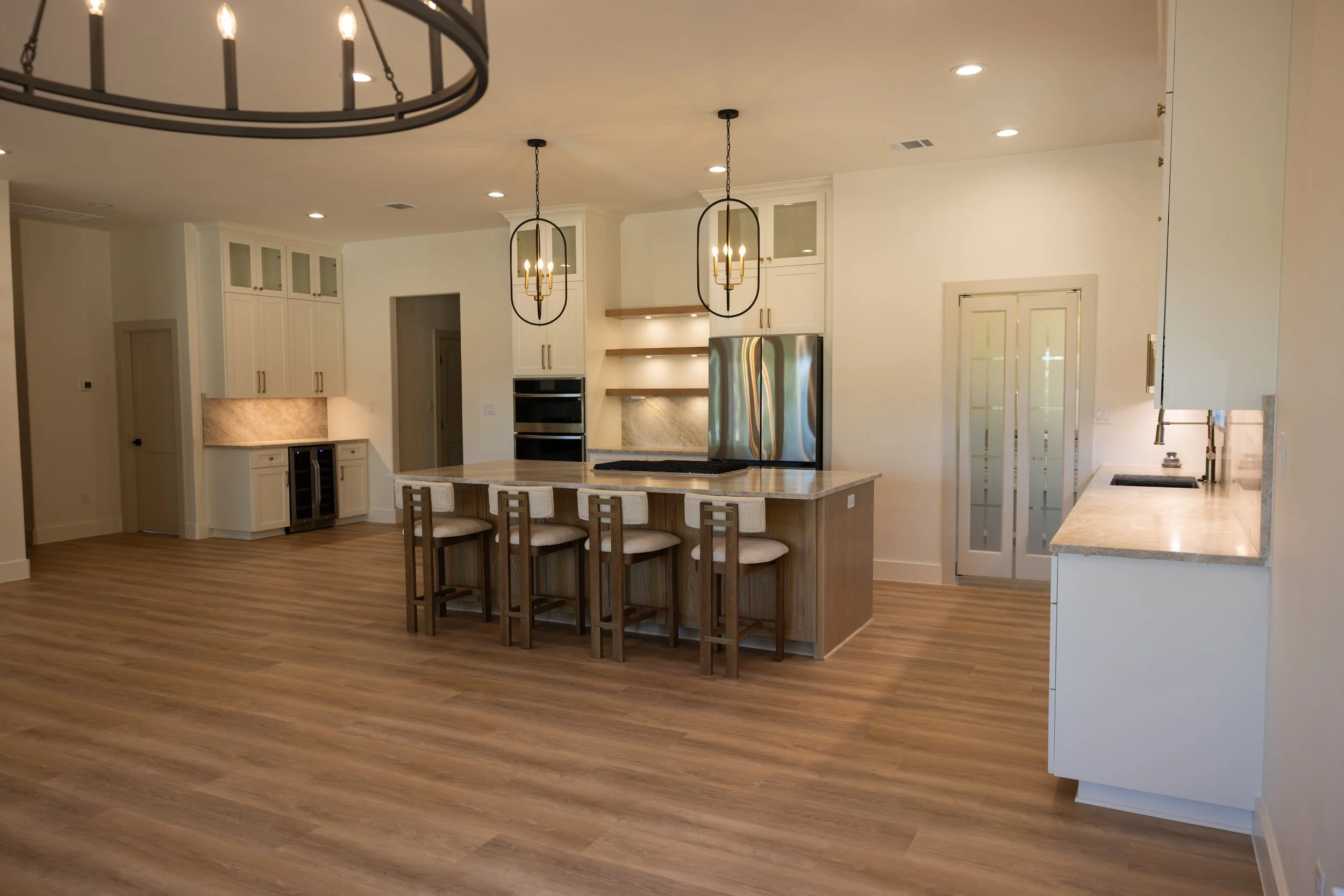 A modern kitchen with an island and bar stools, white cabinets, built-in appliances, pendant lights, and wood flooring.