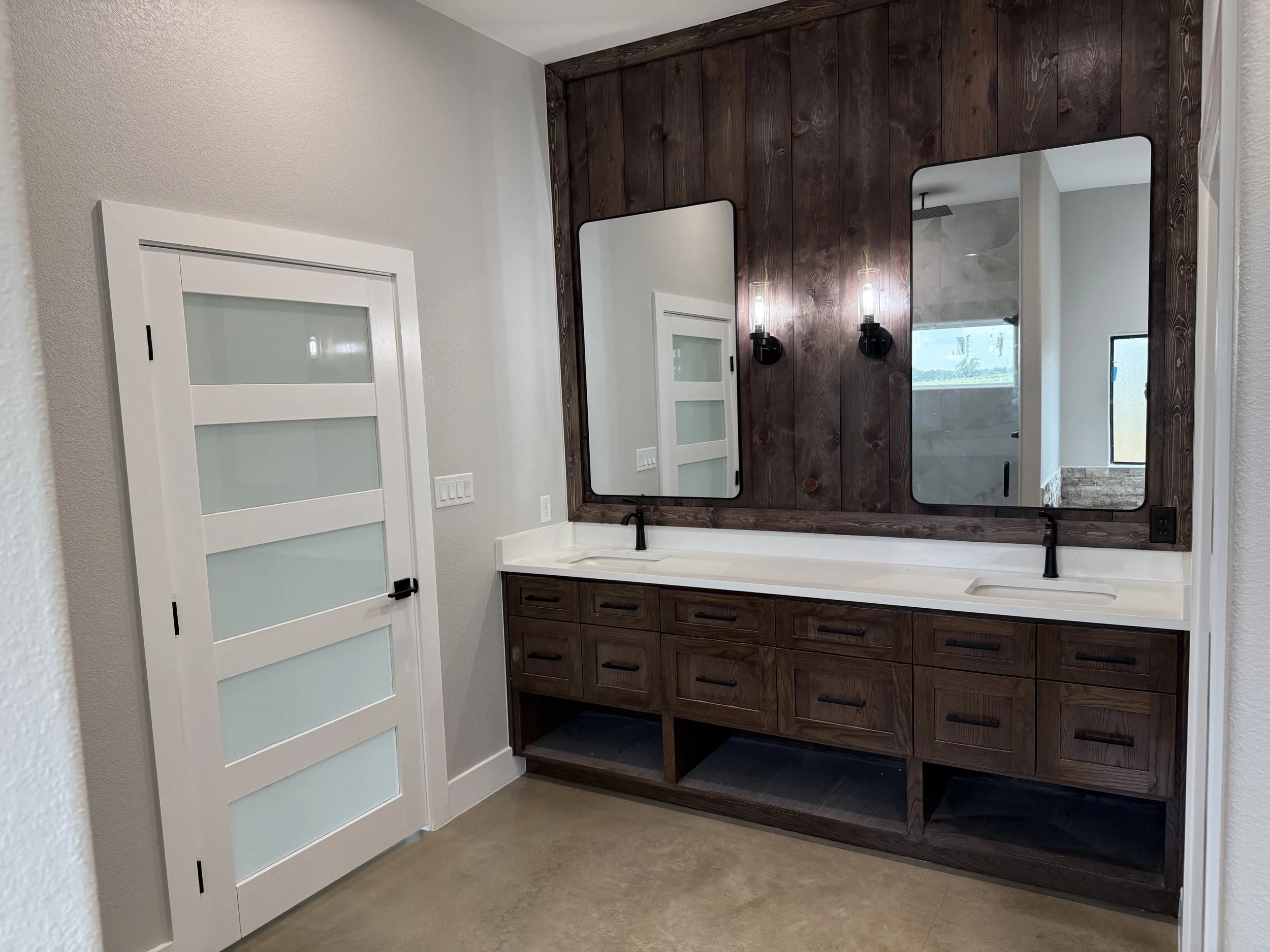Bathroom vanity with two large mirrors, dark wood cabinetry, double sink, black faucet, wall-mounted black light fixtures, and a door with frosted glass panels.