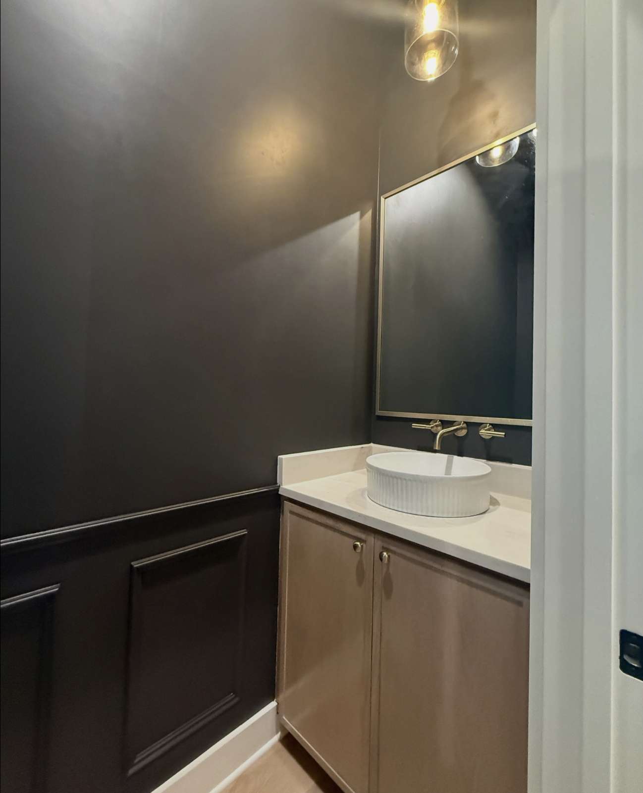 A bathroom vanity with a light-colored cabinet, a white ribbed vessel sink, a gold faucet, and a large rectangular mirror above it. The wall behind the vanity is dark gray.