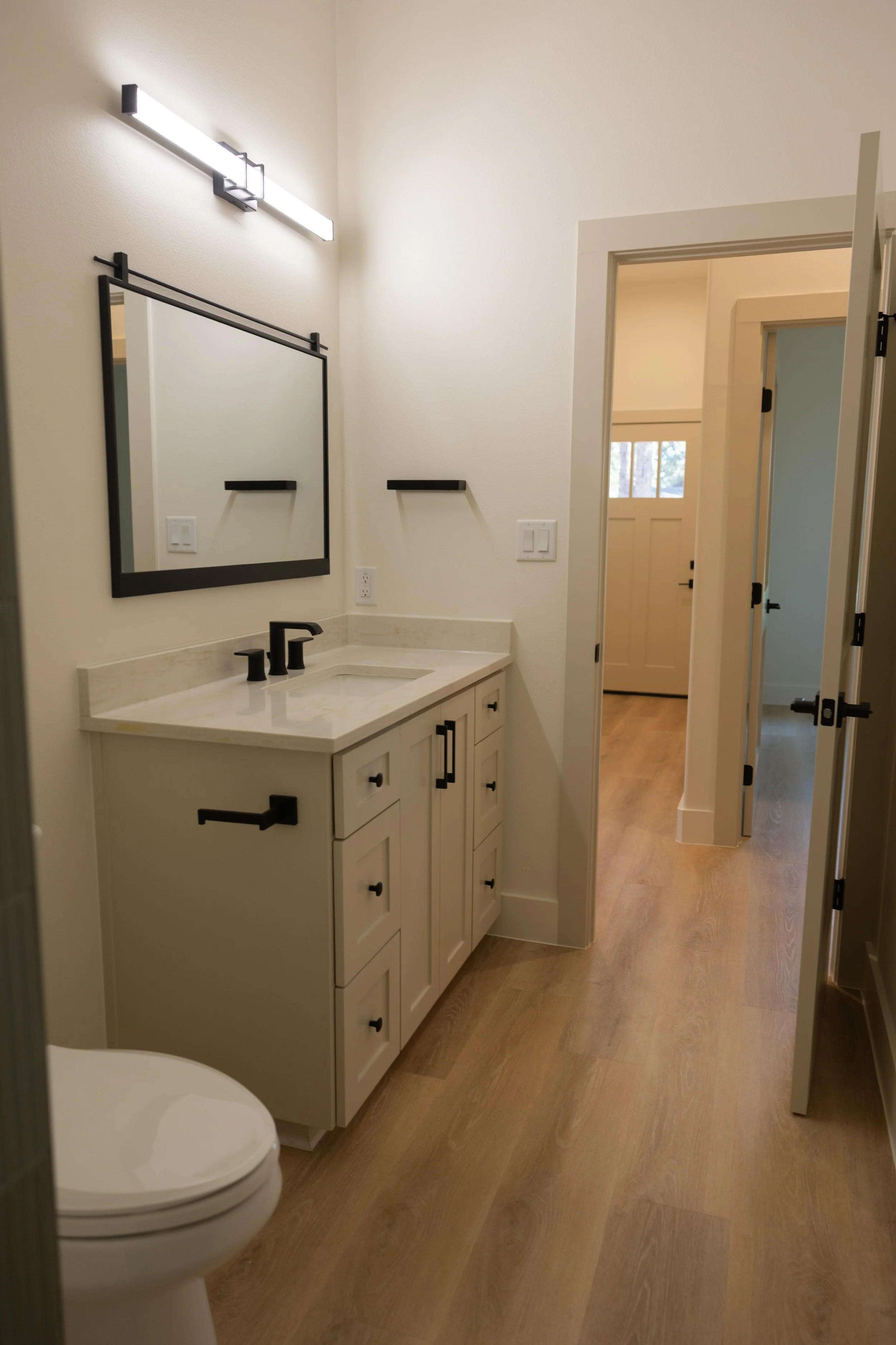 A modern bathroom with a white vanity, black fixtures, a mirror, and a door leading to another room with a window. There is a toilet visible in the lower left corner.