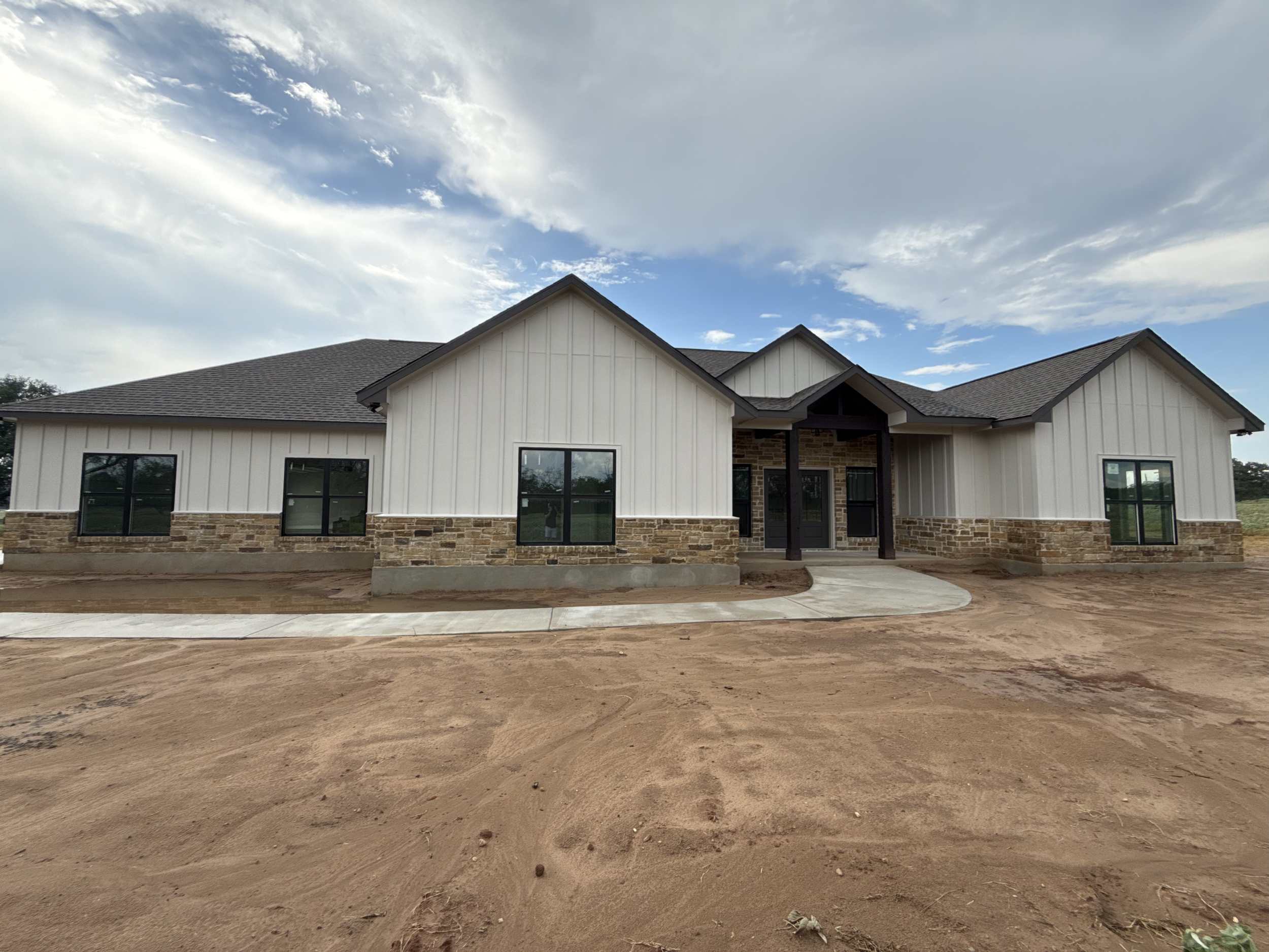 Newly built house with white siding, stone accents at the base, black window frames, and a covered porch, set on a dirt lot with a concrete walkway leading to the front entrance under a partly cloudy sky.