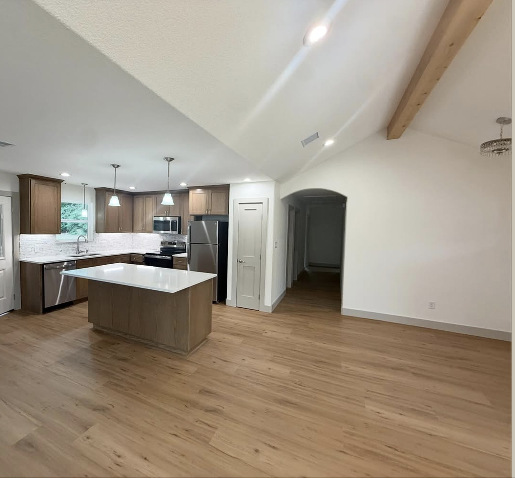 Empty kitchen with wooden floors, white walls, wooden cabinets, stainless steel appliances, a kitchen island with a white countertop, and pendant lights.