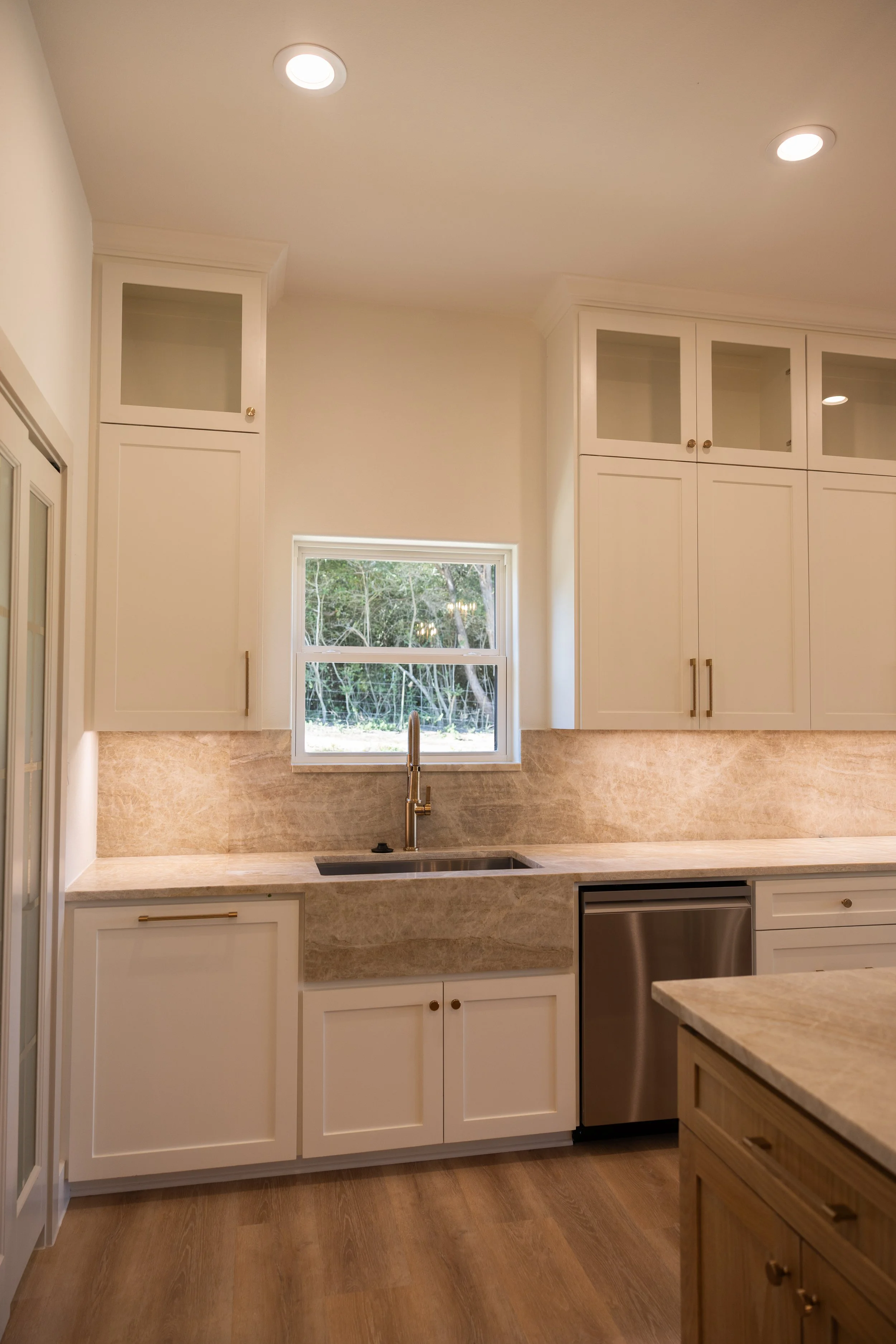 A kitchen with white cabinets, a beige marble countertop, a window above the sink, and wooden flooring.