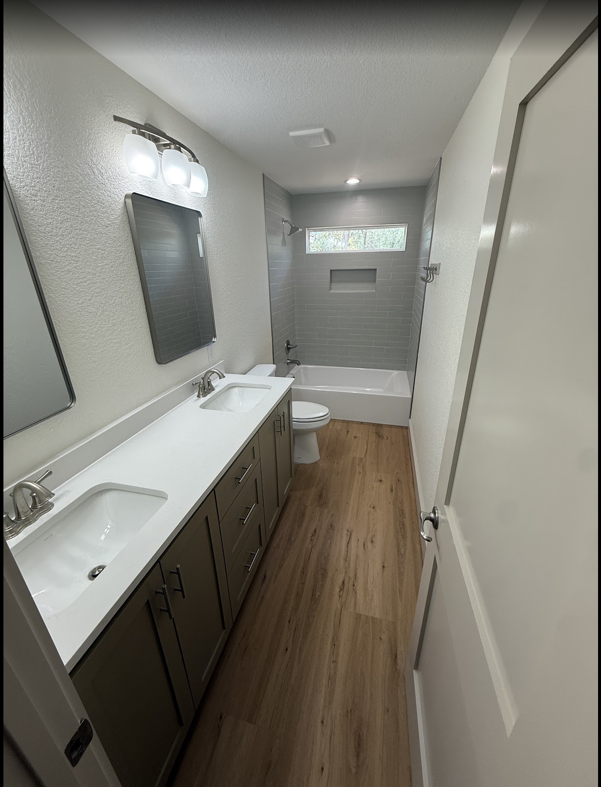 Modern bathroom with dual sinks, a mirror, wall-mounted light fixture, toilet, bathtub with gray tile surround, narrow horizontal window, wood flooring, and white walls.