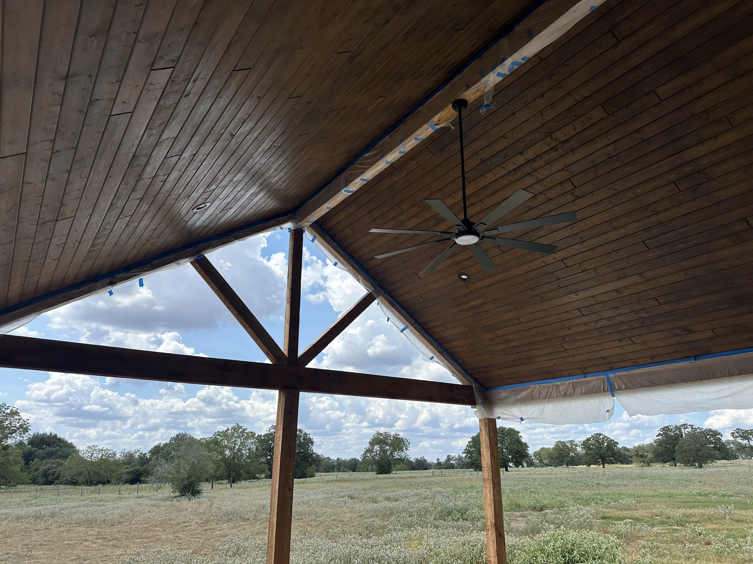 Interior view of a wooden pavilion with a ceiling fan, open sides, and a view of a grassy field with trees and clouds in the sky