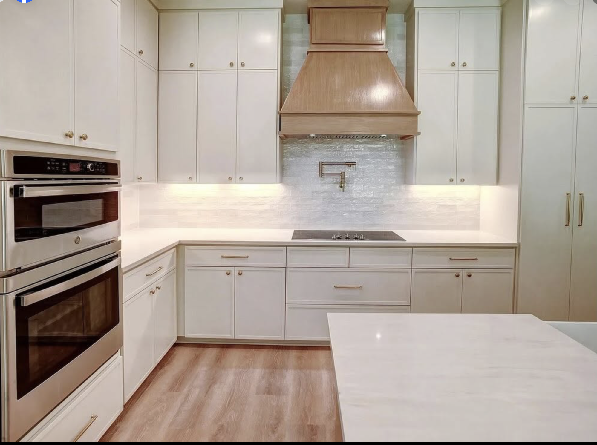 Modern kitchen with white cabinets, wooden accents, stainless steel oven, and wooden range hood, with a wooden floor and light-colored walls.