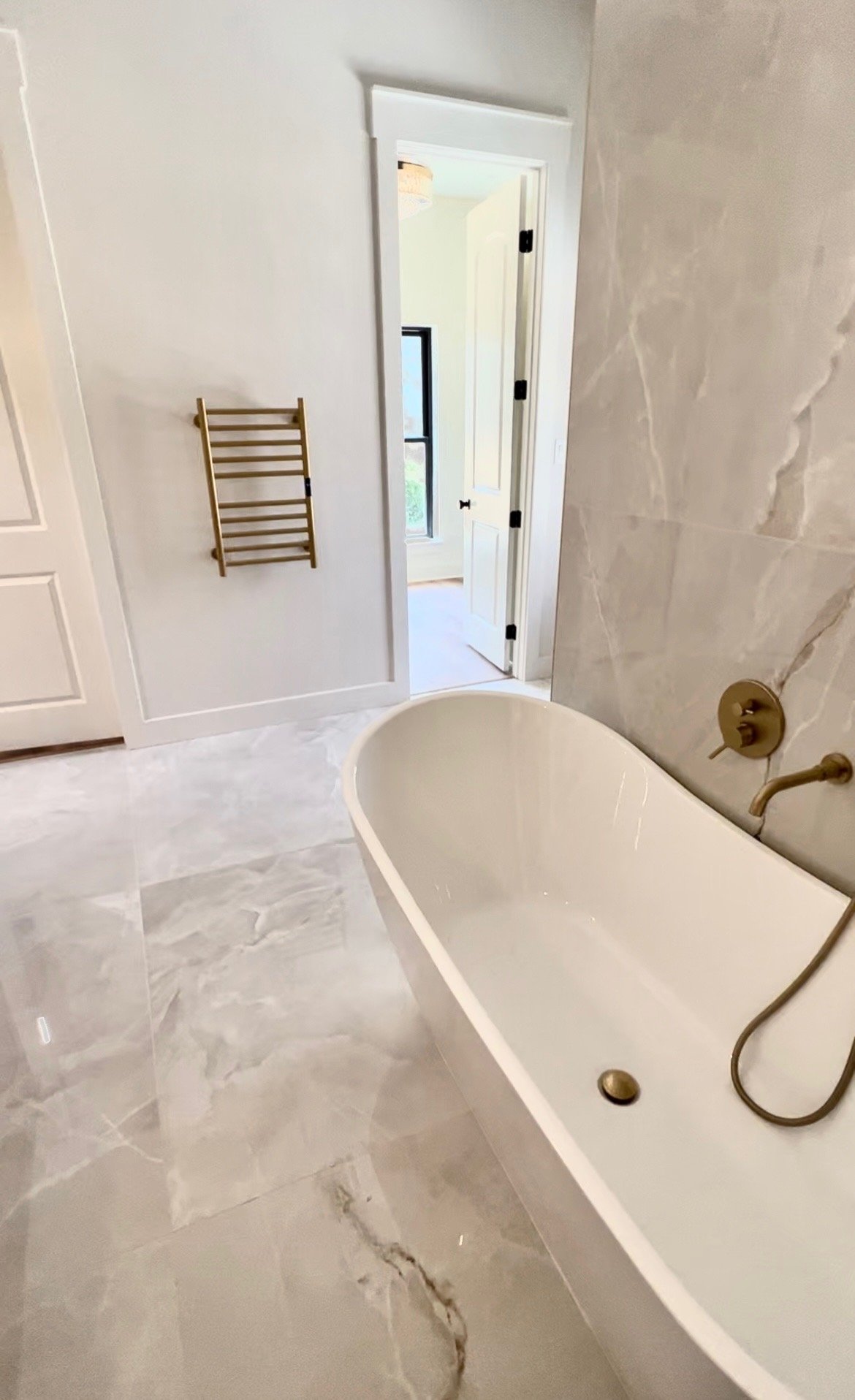 A modern bathroom featuring a freestanding bathtub with brass fixtures, marble-look floor and wall tiles, a wooden towel rack on the wall, and an open door leading to a room with a window.
