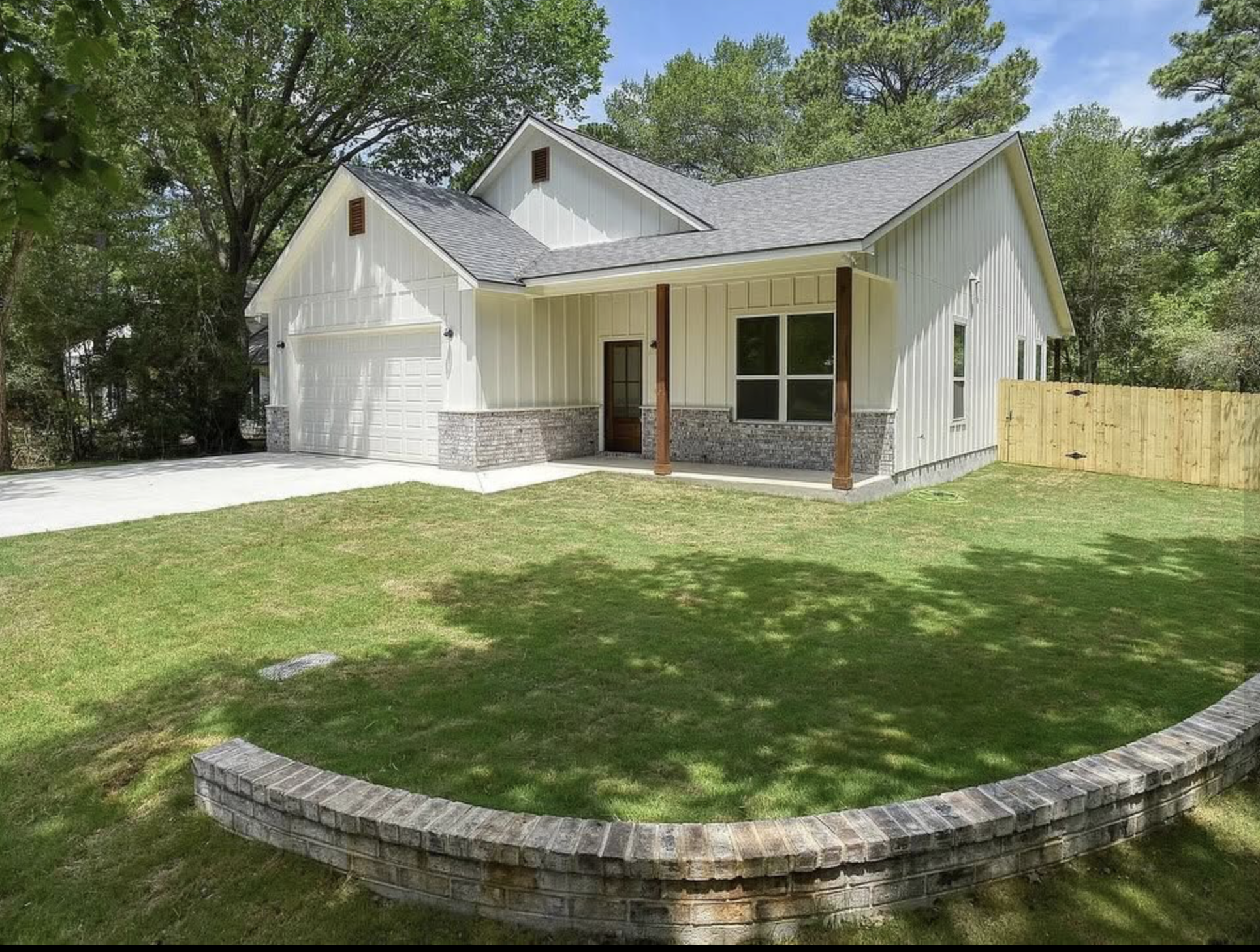 A modern single-story house with a white exterior, a gabled roof, brick accents at the base, a front porch with wooden posts, a garage, and a lush green lawn surrounded by trees and a wooden fence.
