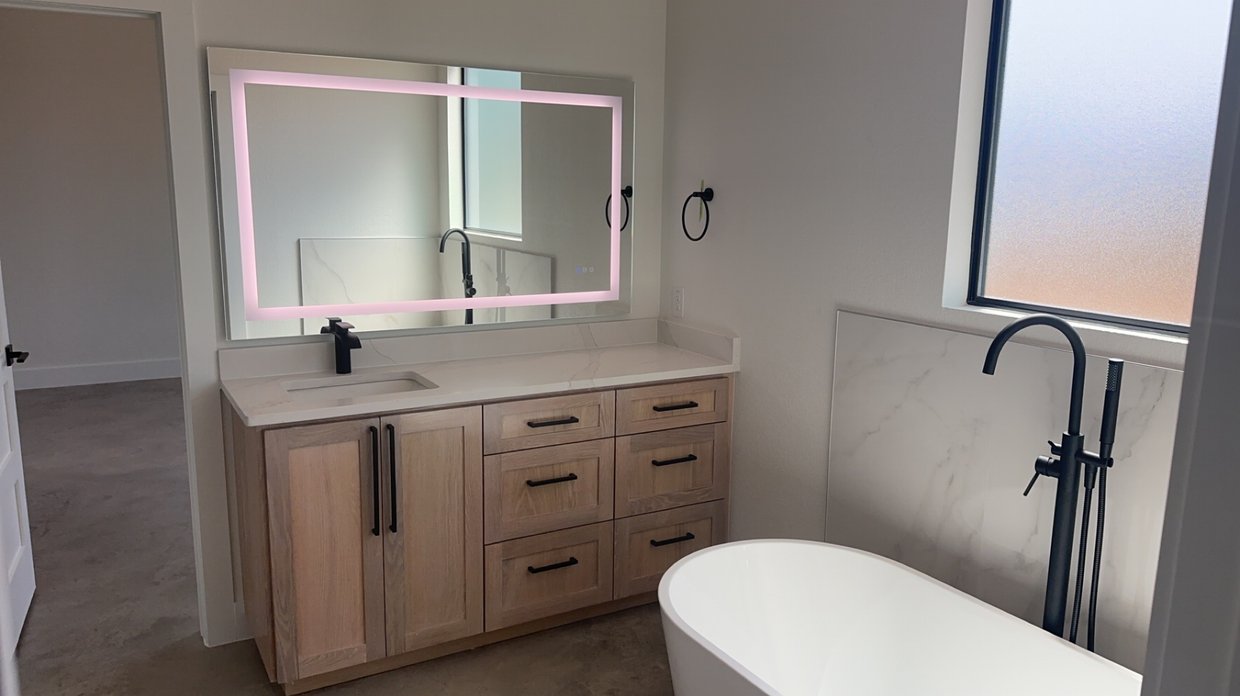 Modern bathroom with wooden vanity, black faucet, large mirror with pink LED backlight, white bathtub, and frosted window.