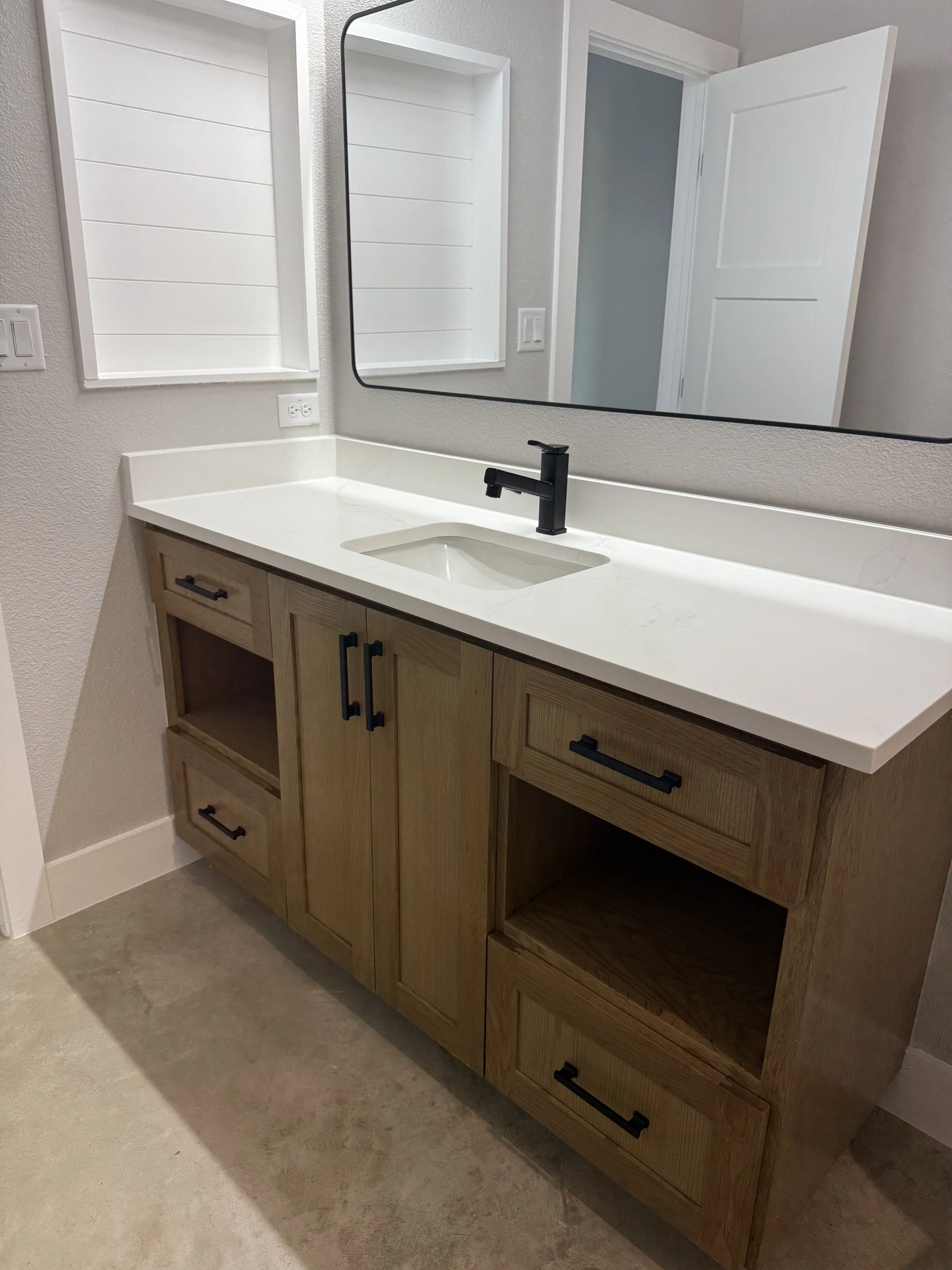 Bathroom vanity with a white countertop, a rectangular sink, black faucet and hardware, wooden cabinetry, a mirror, and a white wall with a window and door.